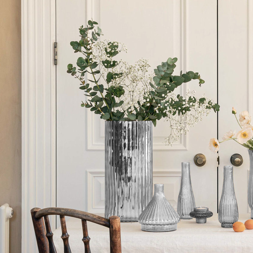 The Ivyline - Daphne Large Ribbed Vase in Clear, topped with greenery and flowers, adds elegance to the table décor beside a white paneled door and a wooden chair.