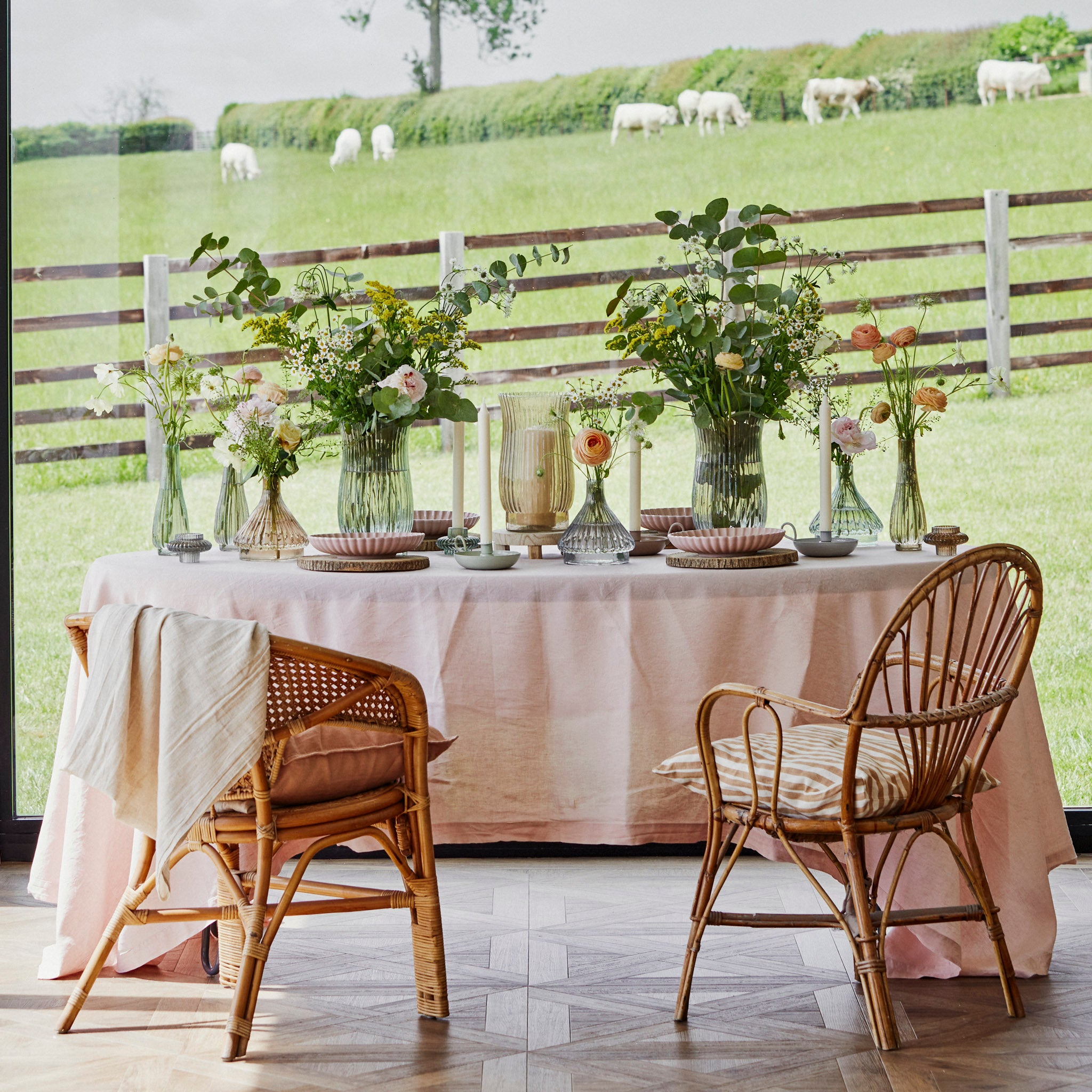 The elegant table, featuring the Ivyline - Daphne Large Ribbed Vase in Sage filled with flowers, sets the stage for sustainable living while overlooking a green pasture with grazing sheep through large windows.
