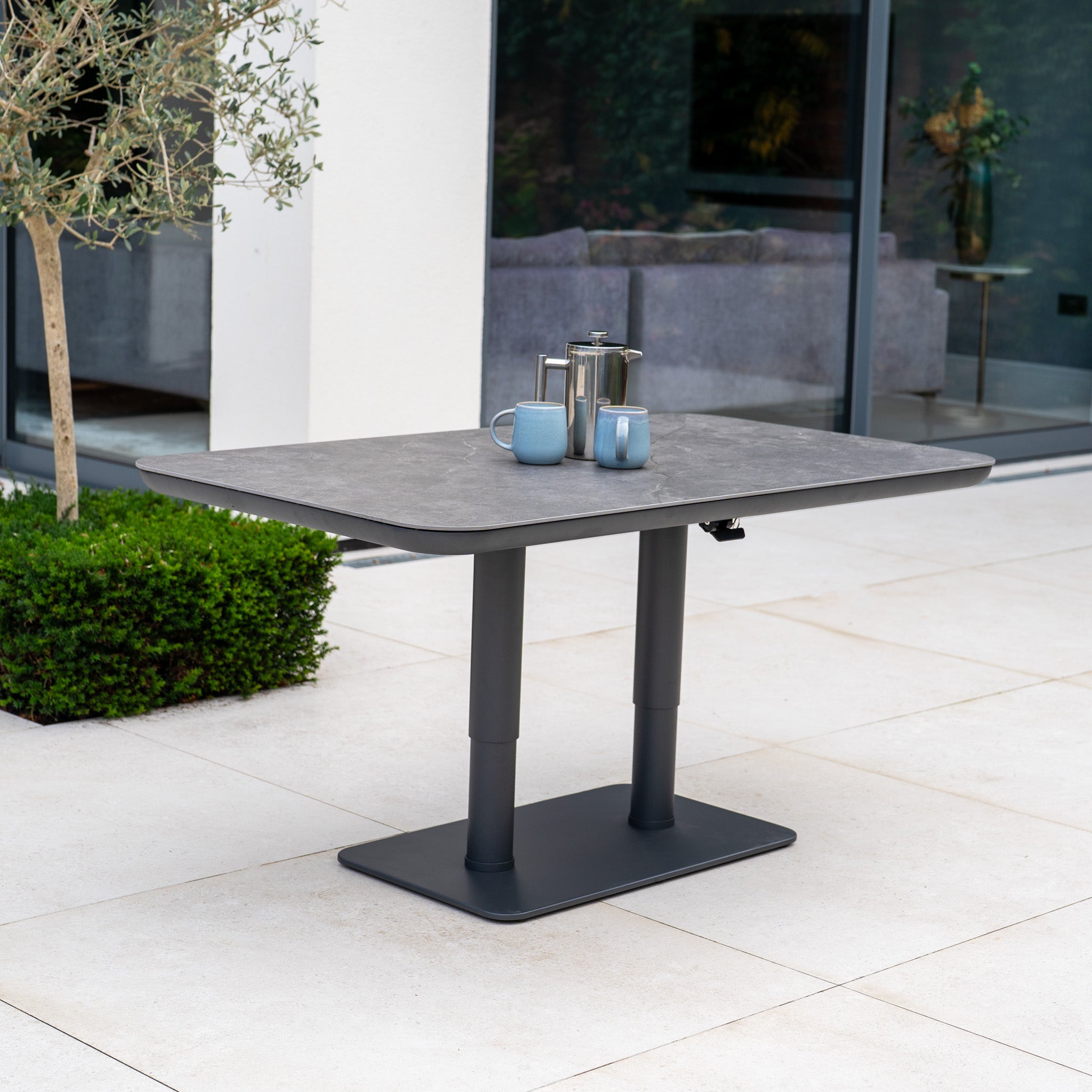 A modern outdoor table with a coffee pot and two mugs sits on a tiled patio near greenery, perfectly complementing the Luna Outdoor Fabric Curved U-Shape Sofa Set with Altura Rising Table in Oyster Grey.