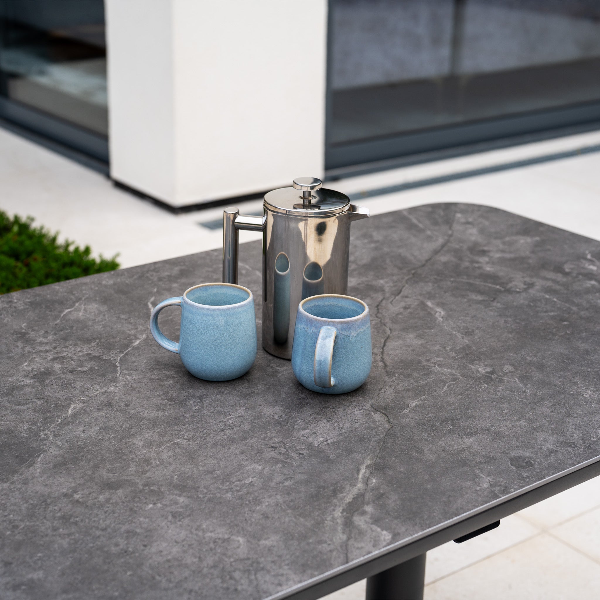 A metal French press and two blue mugs rest on the Altura Rising Table in Grey, part of the Luna U-Shape Outdoor Fabric Sofa Set featuring a durable aluminium frame, near a modern building.