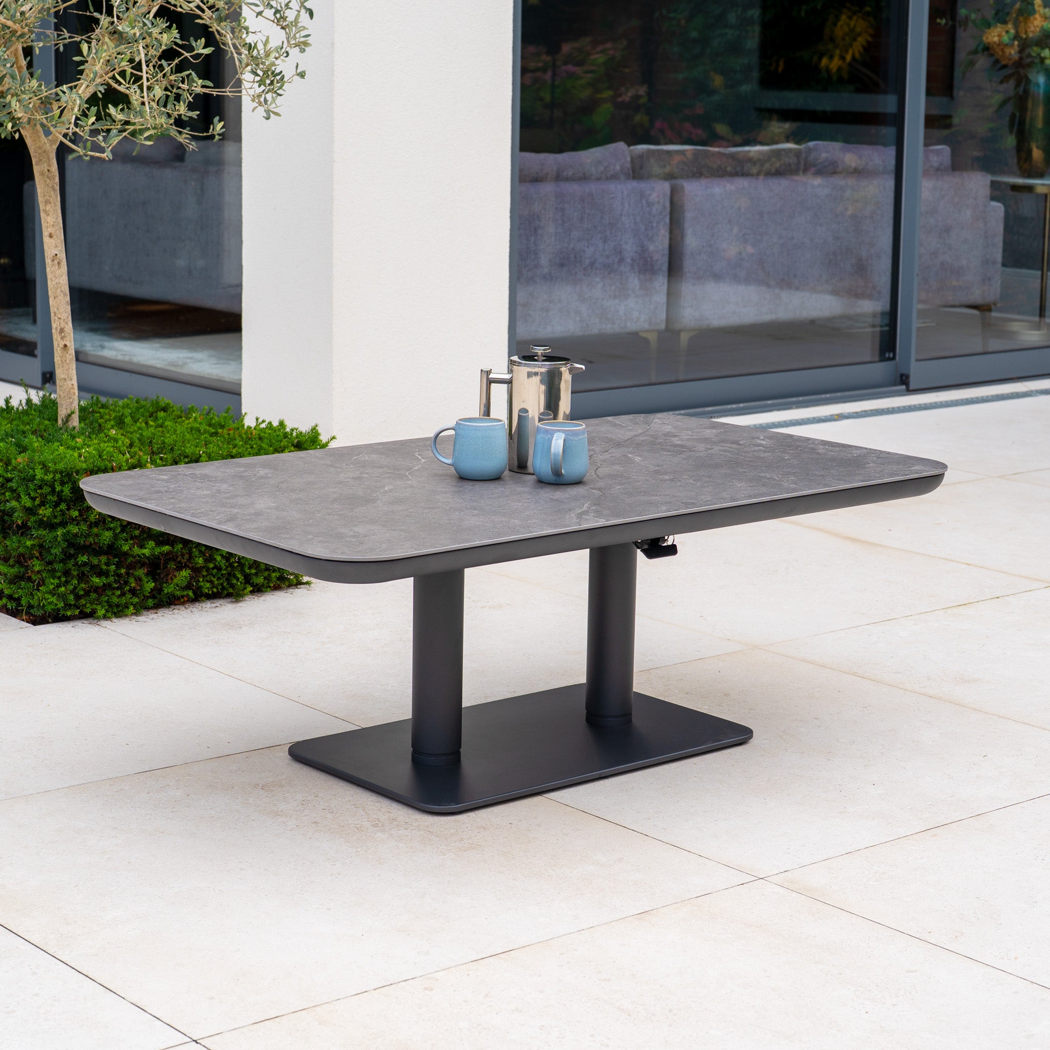 Modern outdoor table with mugs and a coffee press on a patio beside large glass doors, perfectly complementing your garden furniture or the Pello Deluxe Curved Sofa Set with Altura Rising Table in Grey for stylish outdoor living.