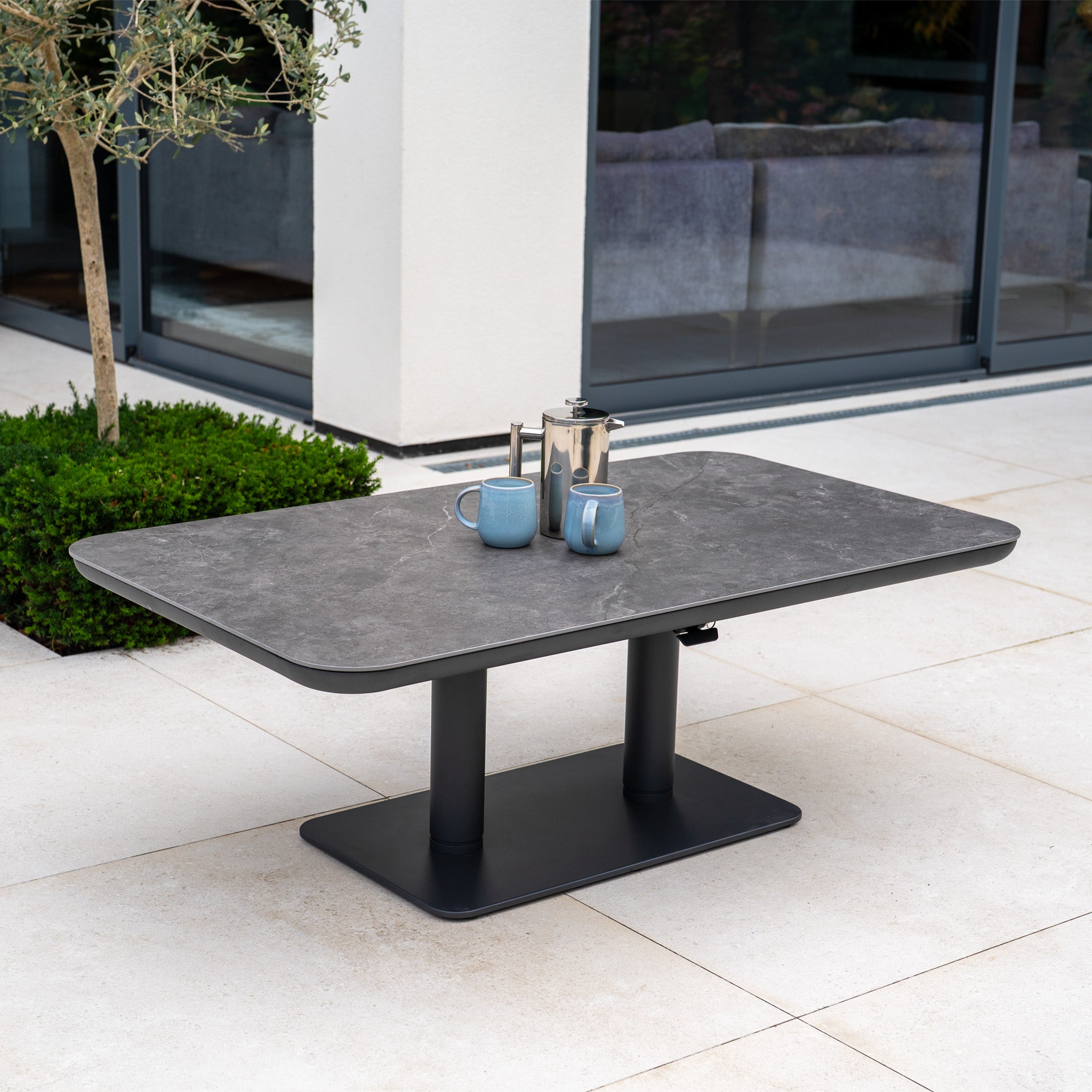 The Luna Outdoor Fabric Curved U-Shape Sofa Set with Altura Rising Table in Oyster Grey, with mugs and a coffee pot on the table, sits on a patio near a window and green plants for stylish outdoor comfort in any season.
