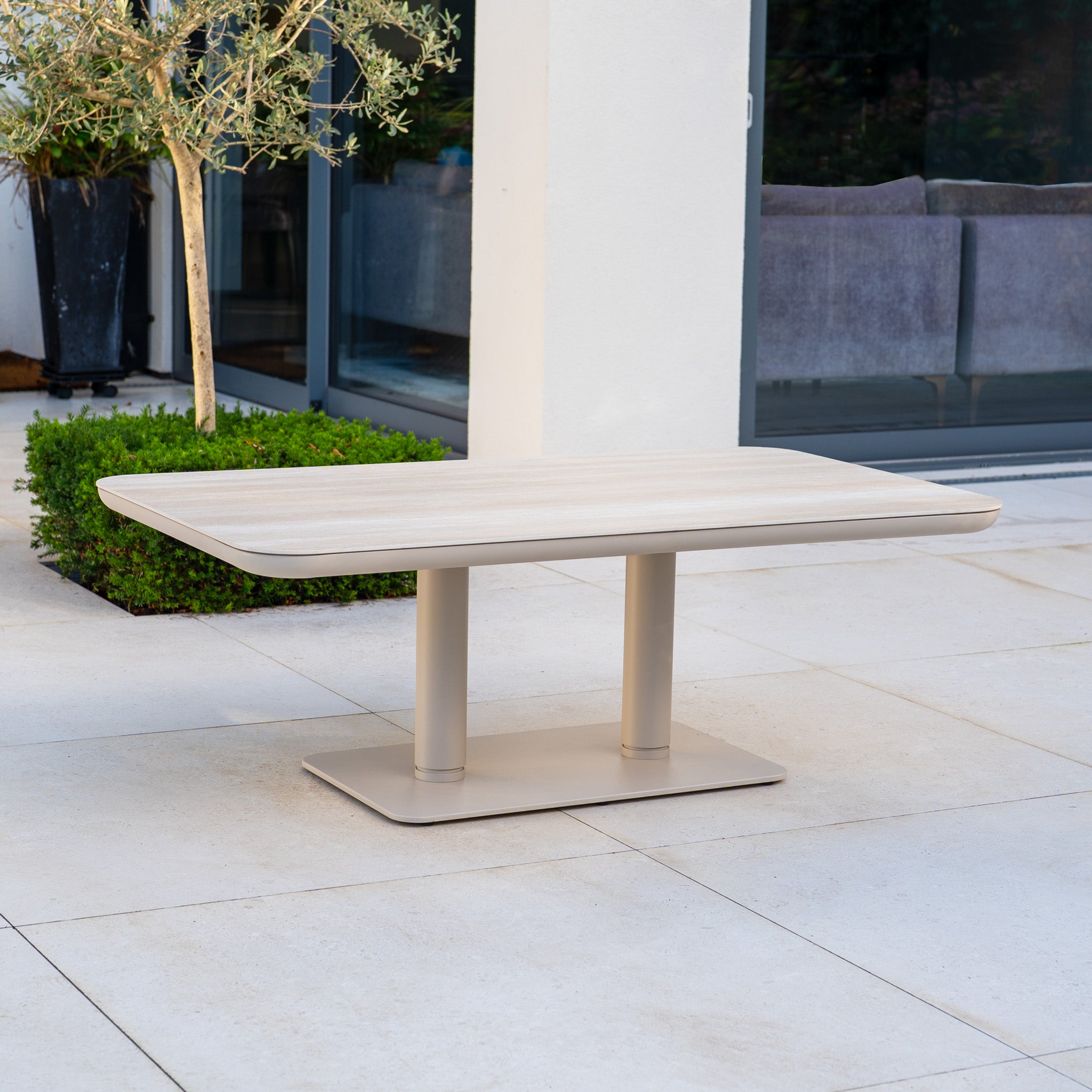 The Dekota Large Corner Sofa Dining Set with Altura Rising Table in Fawn features a modern rectangular table on a tiled patio by glass doors, complementing your outdoor space for relaxed gatherings.