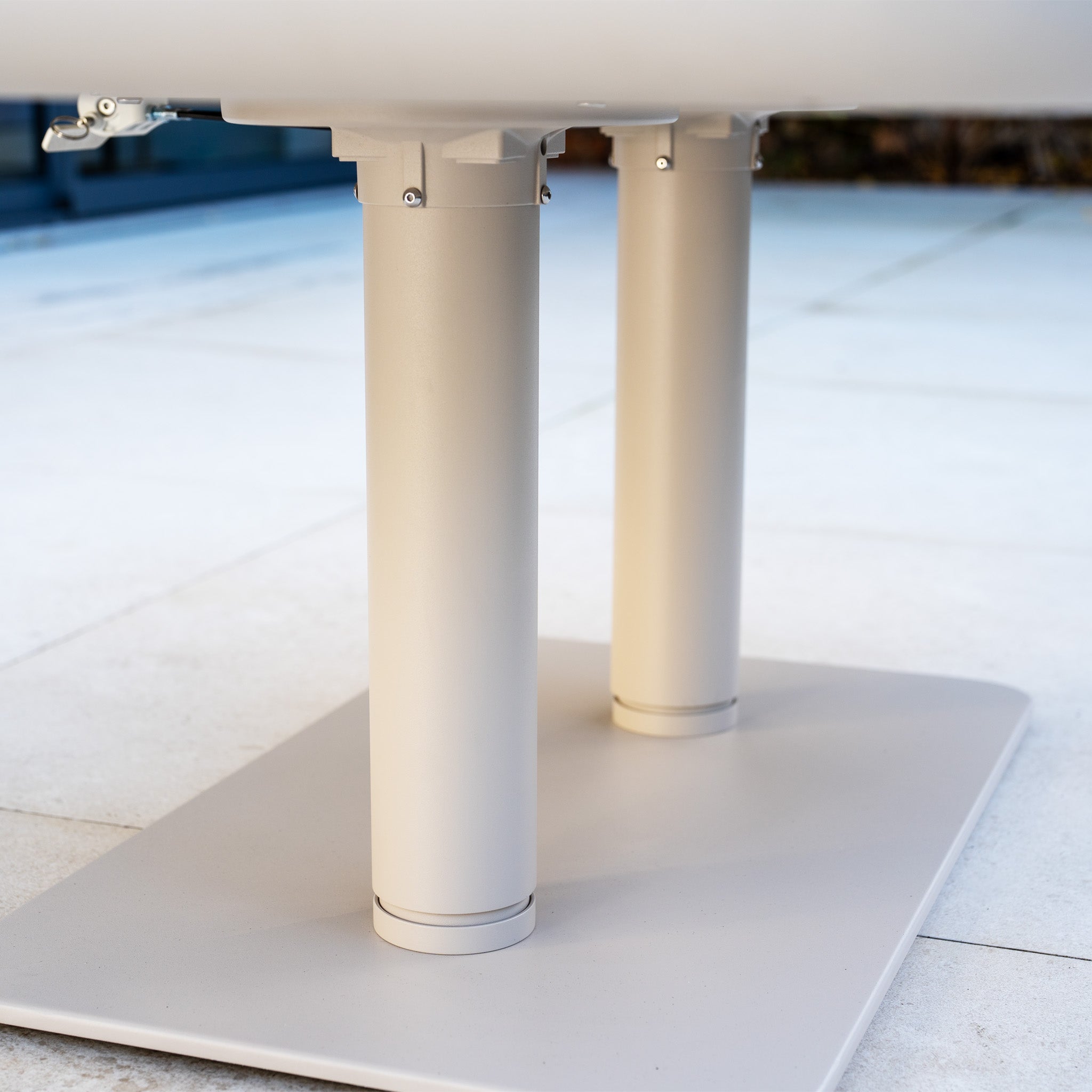 Close-up view of two cylindrical metal table legs attached to a flat rectangular base on a tiled floor, part of the Alva Corner Dining Set with Rising Table in Fawn (Right Hand)—a refined pick for contemporary garden spaces.