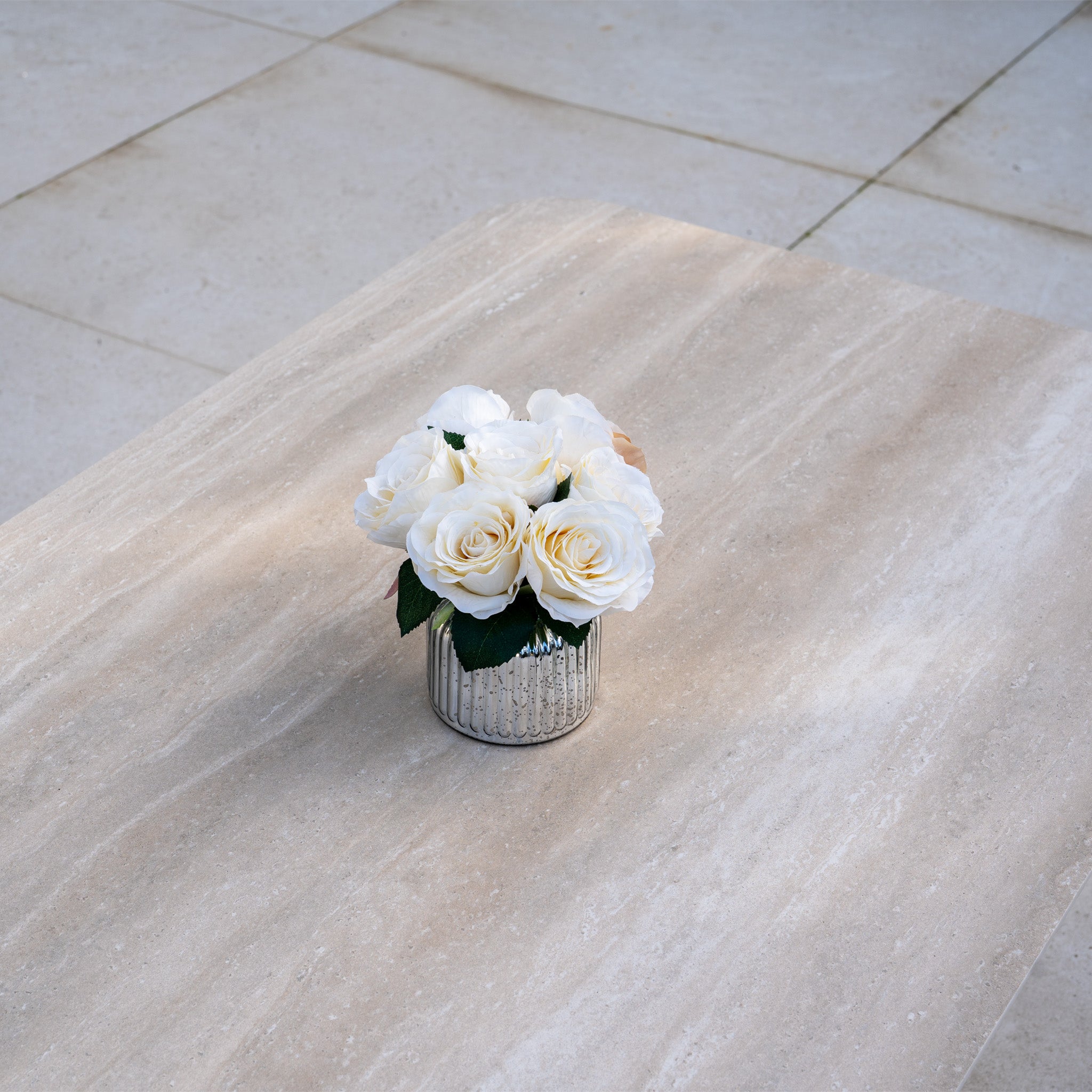 A small glass vase with white roses sits on the Altura Rising Table of the Pello Deluxe Curved Sofa Set in Fawn, featuring quick dry garden cushions, set against a tiled floor background.