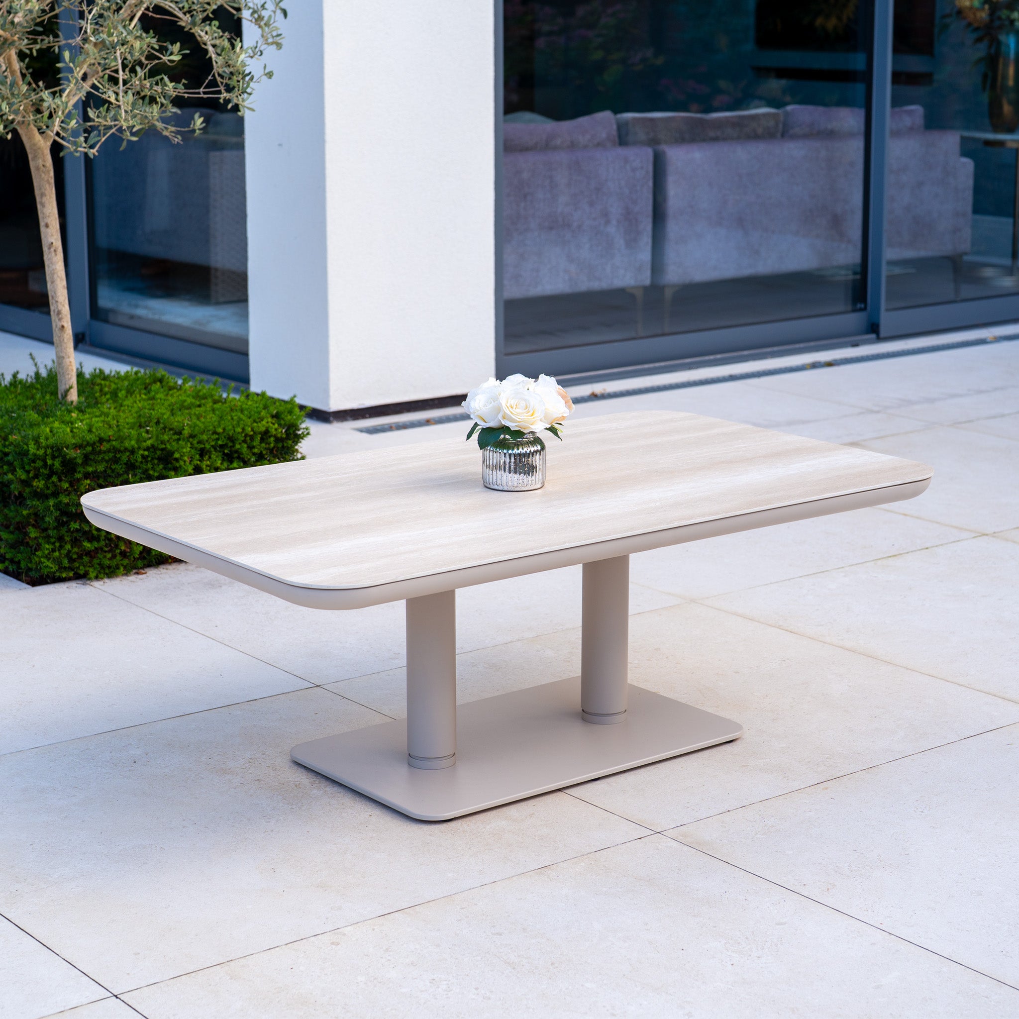 The Luna Outdoor Fabric Large Corner Sofa Set with Altura Rising Table in Fawn is arranged on a tiled patio near a glass door, creating a modern, weather-resistant space ideal for relaxing outdoors.