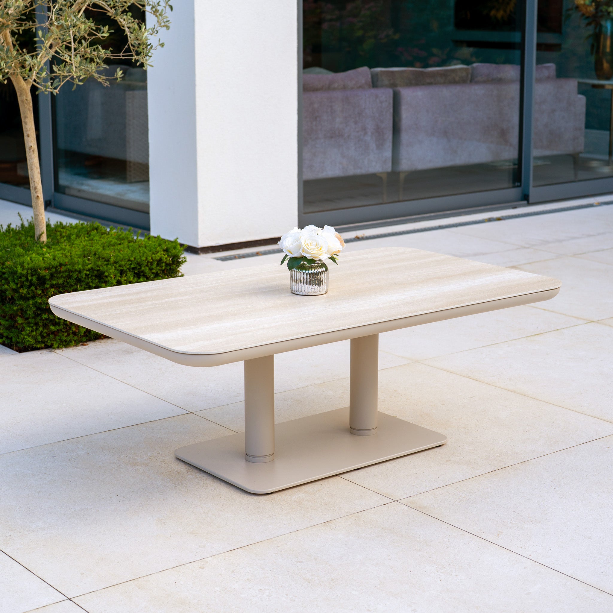 The Savona Luxury Outdoor Large Corner Sofa Set with Altura Rising Table in Oatmeal sits on a tiled patio near glass doors and greenery, styled with a vase of white flowers.