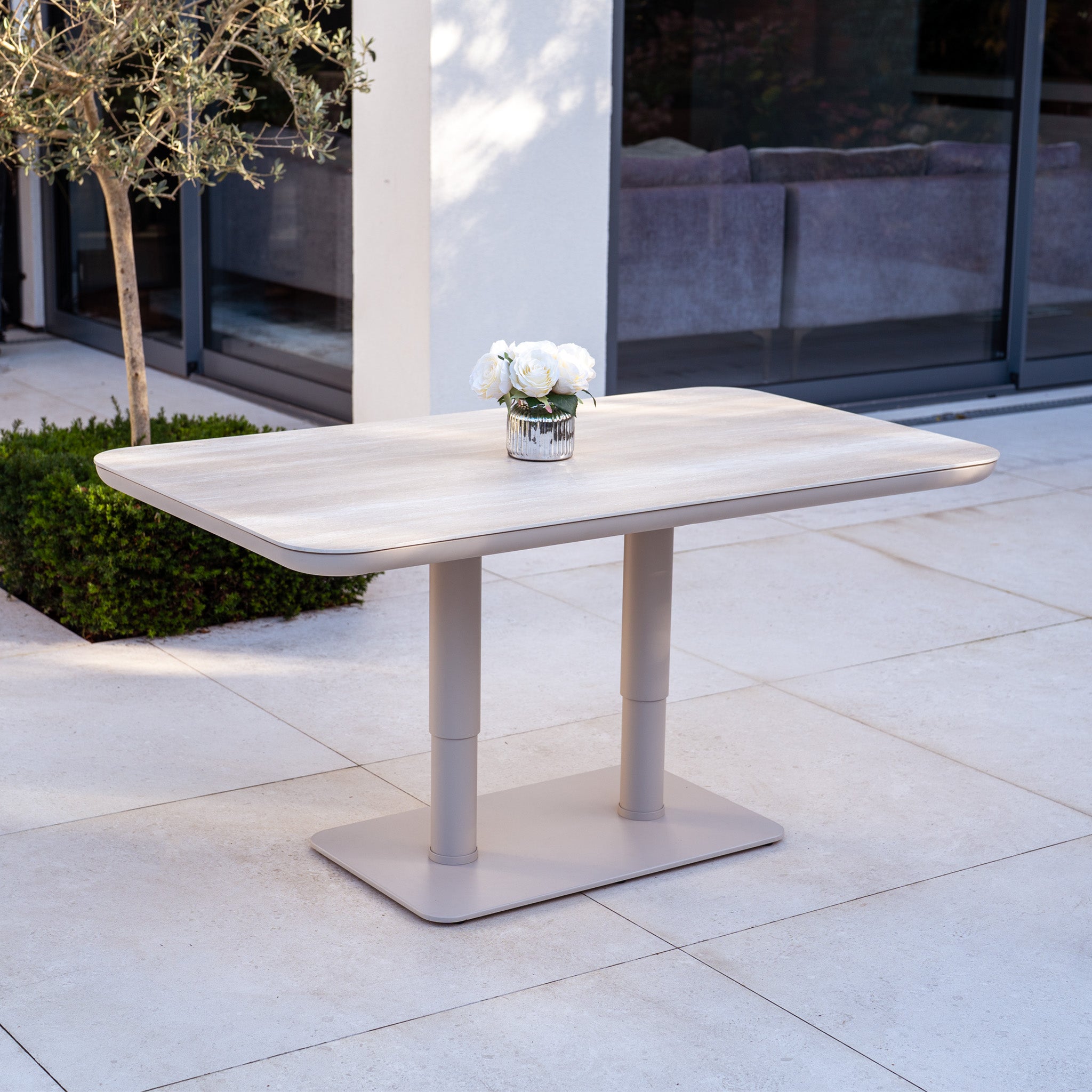 The Luna Outdoor Fabric Large Curved Corner Sofa Set with Altura Rising Table in Fawn creates an inviting all-weather setup, displayed with a small vase of white flowers on a tiled patio near a modern building.