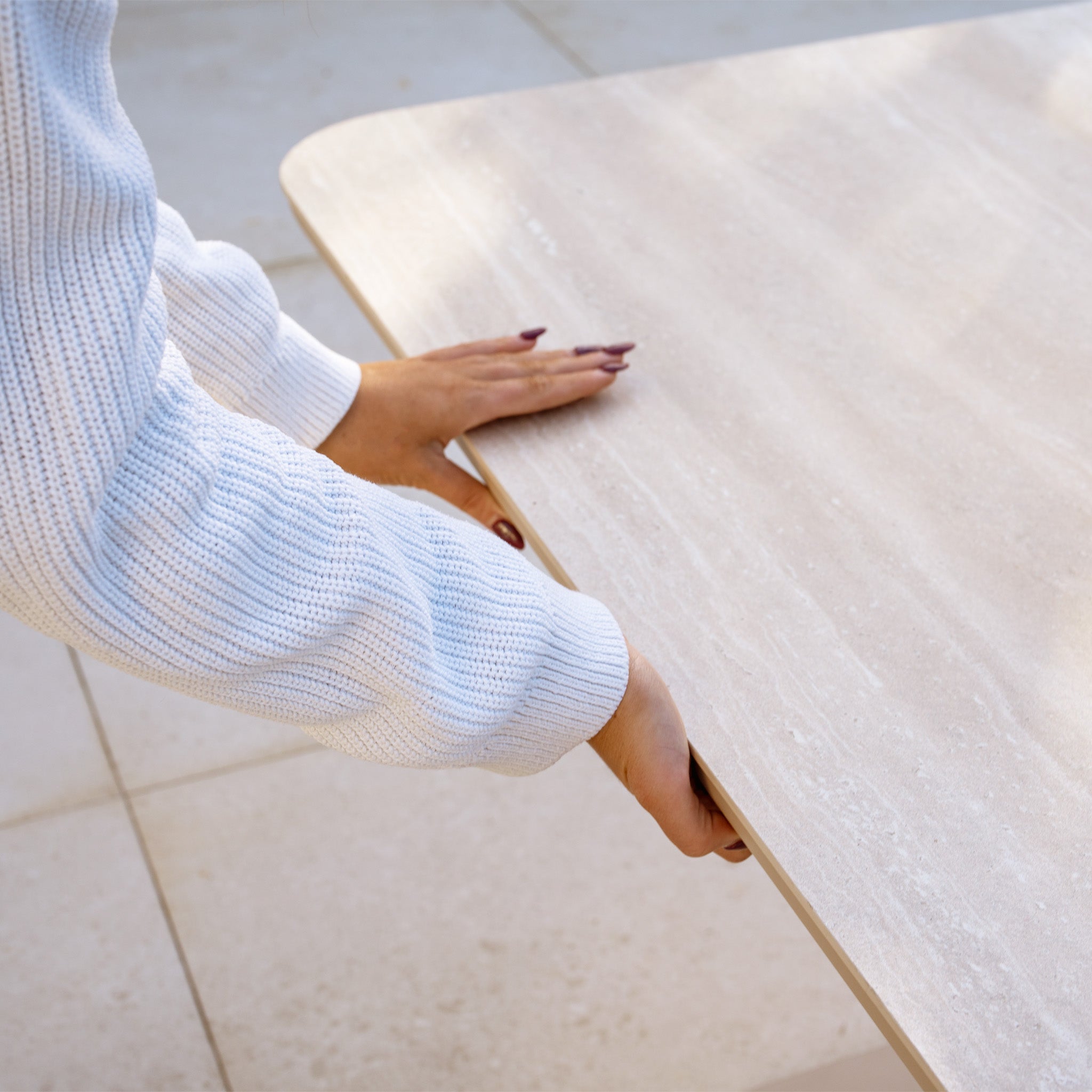 A person in a white sweater holds the edge of an Altura Rising Table beside a Como Corner Sofa Set with Altura Rising Table in Oatmeal, both placed on a tiled floor.