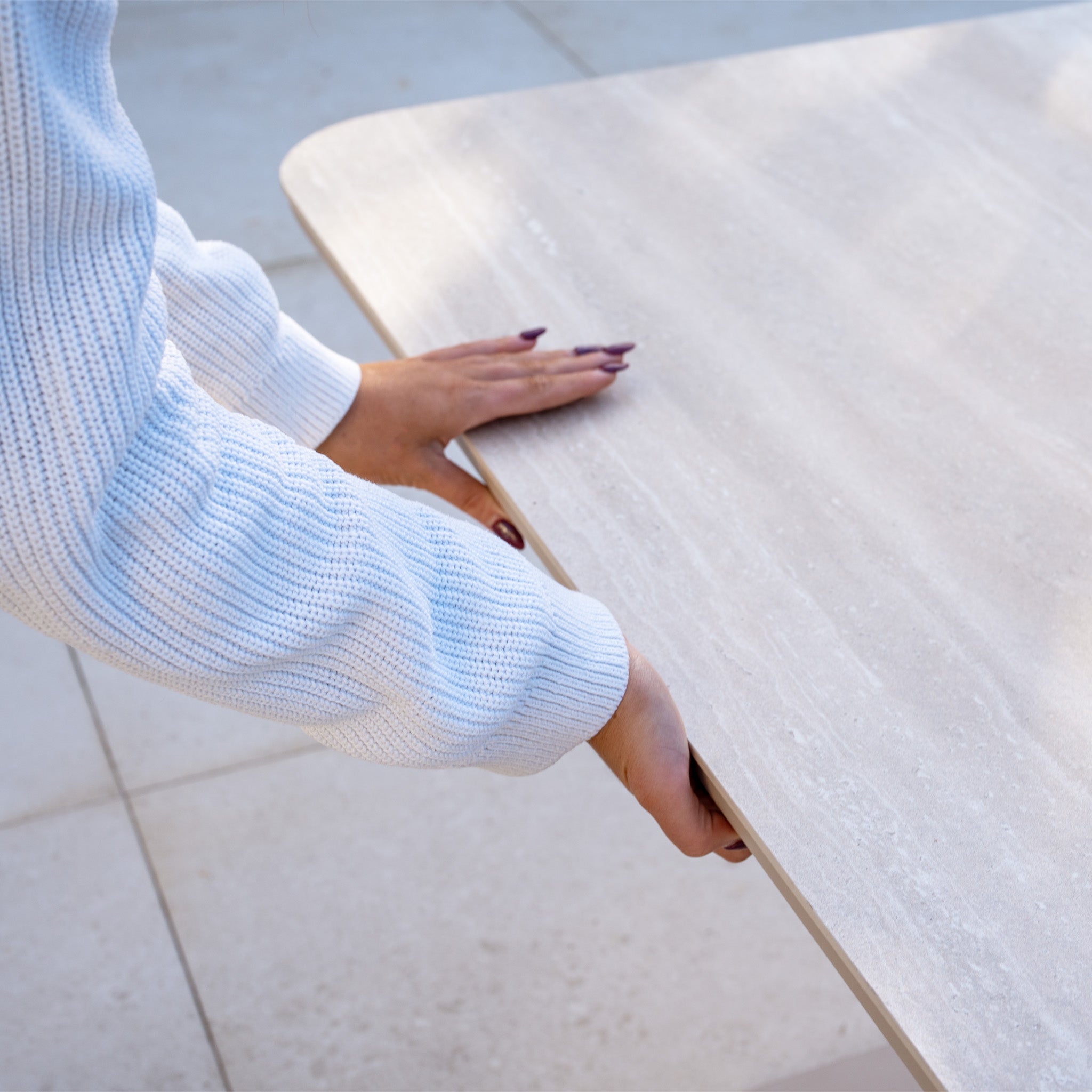 Person in a white sweater holding the Altura Rising Table from the Luna U-Shape Outdoor Fabric Sofa Set in Fawn, ideal with weather-resistant garden furniture on a tiled floor.