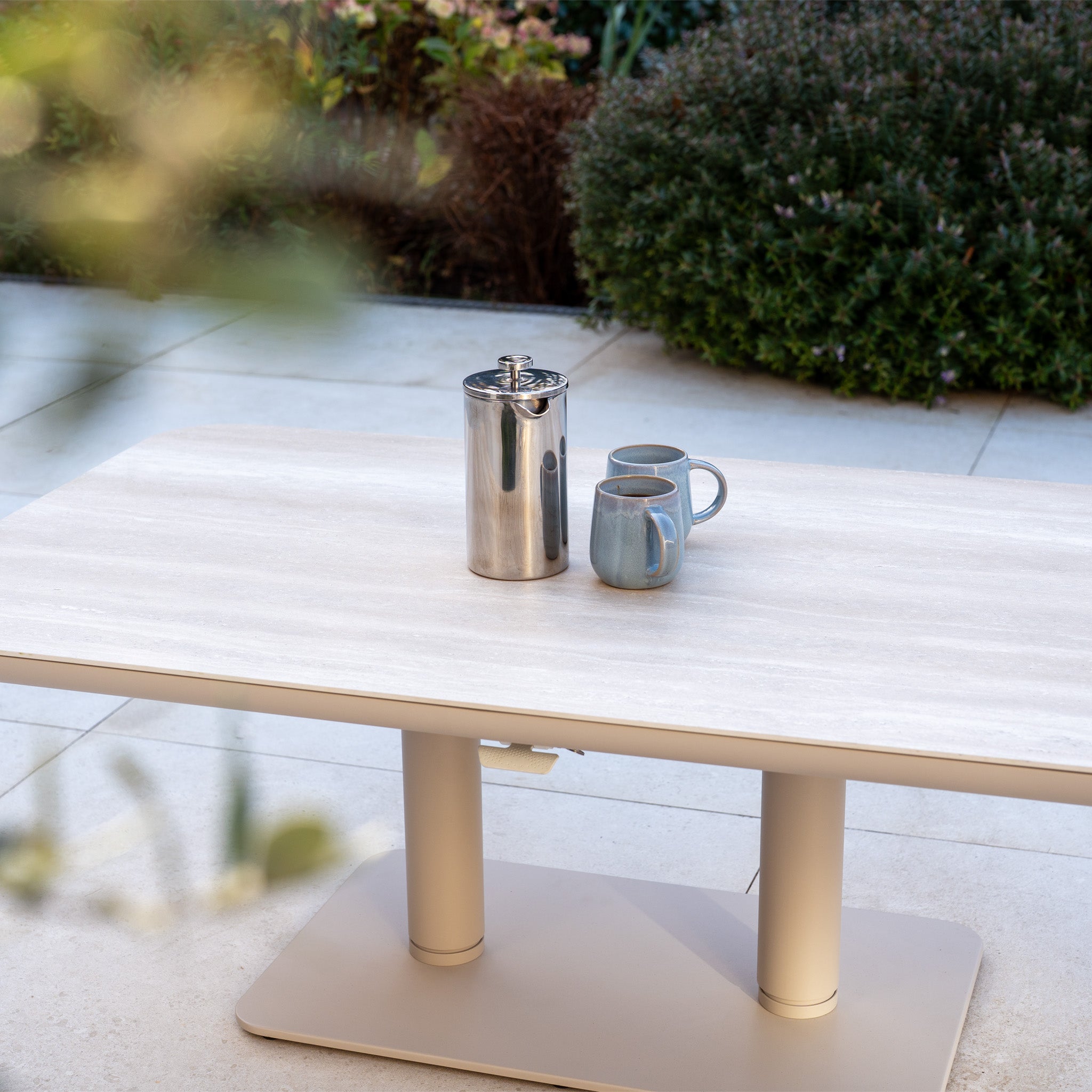 A Luna U-Shape Outdoor Fabric Sofa Set with an Altura Rising Table in Fawn, holding a metal coffee pot and two mugs, is featured as part of weather-resistant garden furniture in a serene garden setting.