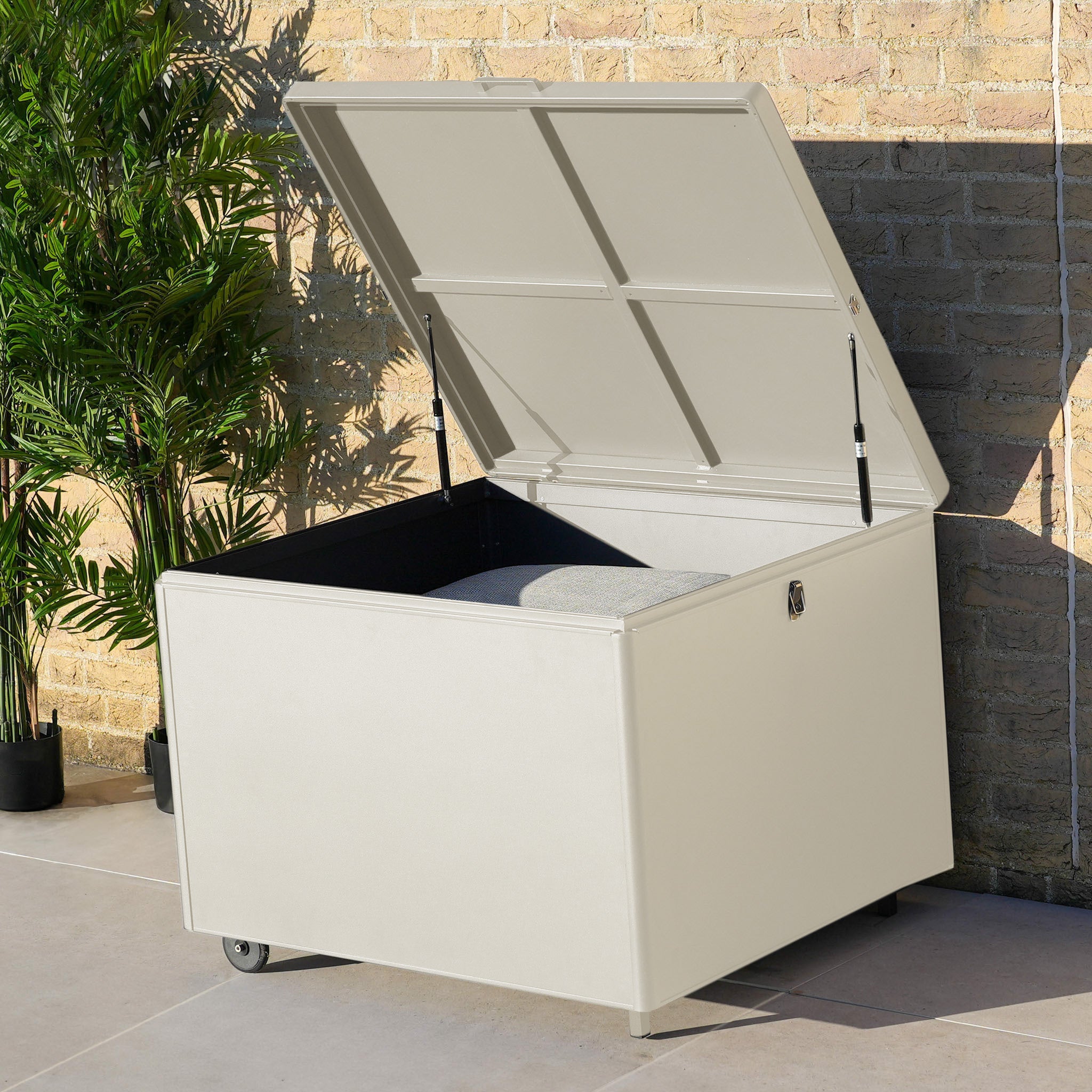 A Small Aluminium Storage Box in Latte with an open lid sits on a patio near a brick wall and potted plants, providing practical outdoor storage while blending into your garden space.