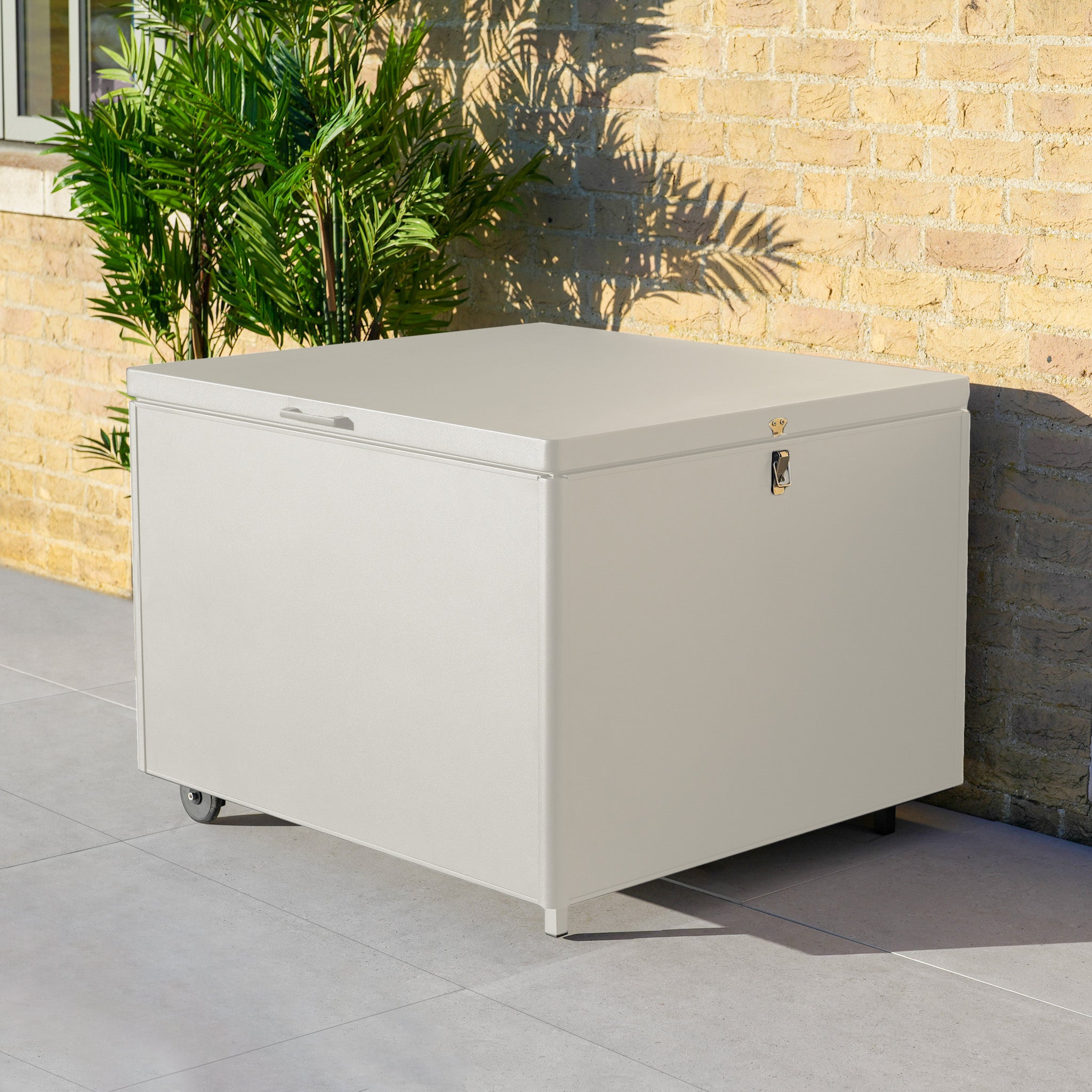 Small Aluminium Storage Box in Latte—ideal for watertight garden storage—sits next to a brick wall and a green potted plant.
