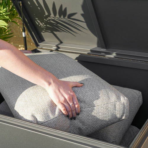 Person placing a gray cushion inside a Small Aluminium Storage Box in Grey with a watertight seal, surrounded by plants in a modern garden.
