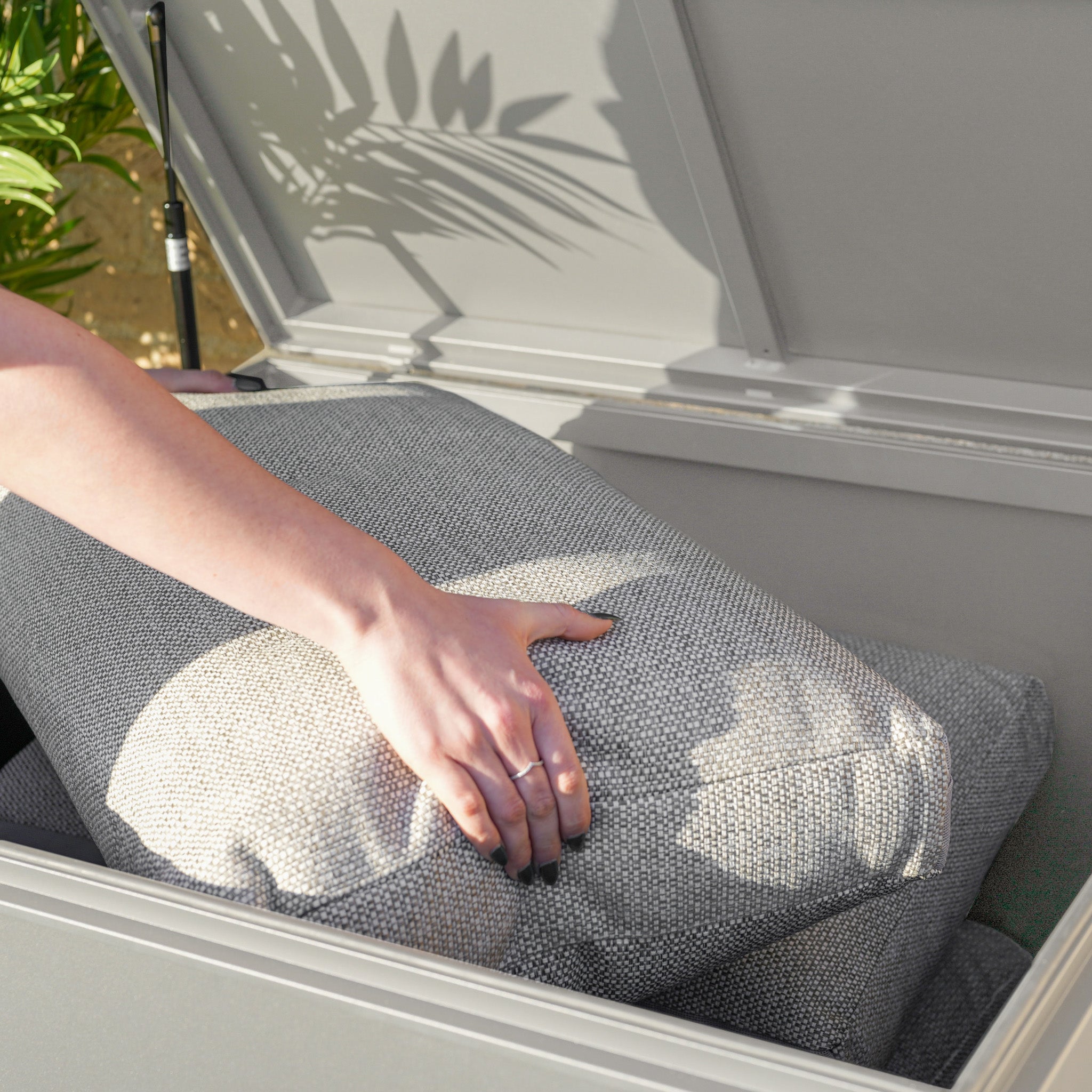 A person places a gray cushion inside the Small Aluminium Storage Box in Latte with the lid open, ensuring their belongings stay protected in this watertight garden box.