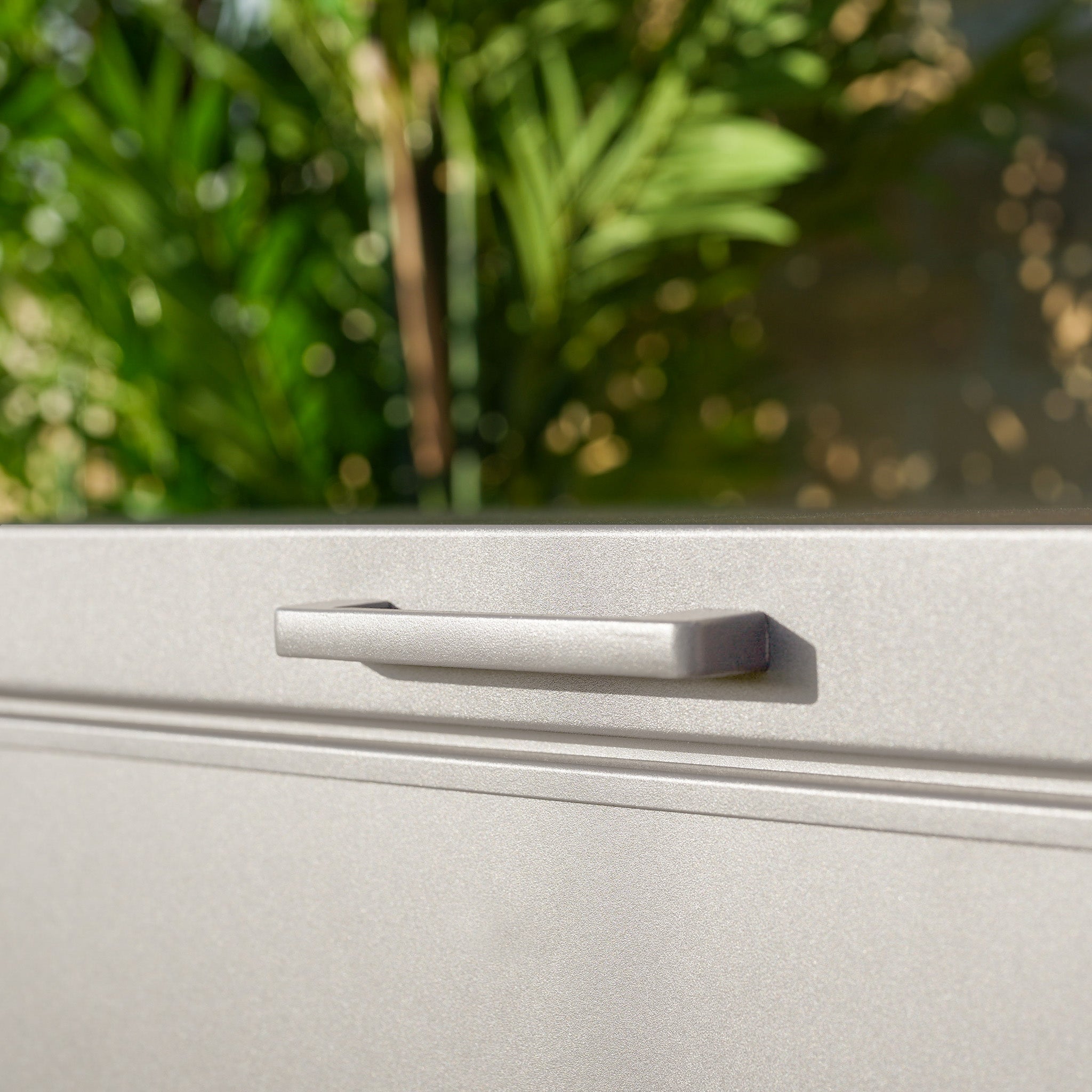 Close-up of the metallic handle on the Small Aluminium Storage Box in Latte, with blurred green plants in the background.