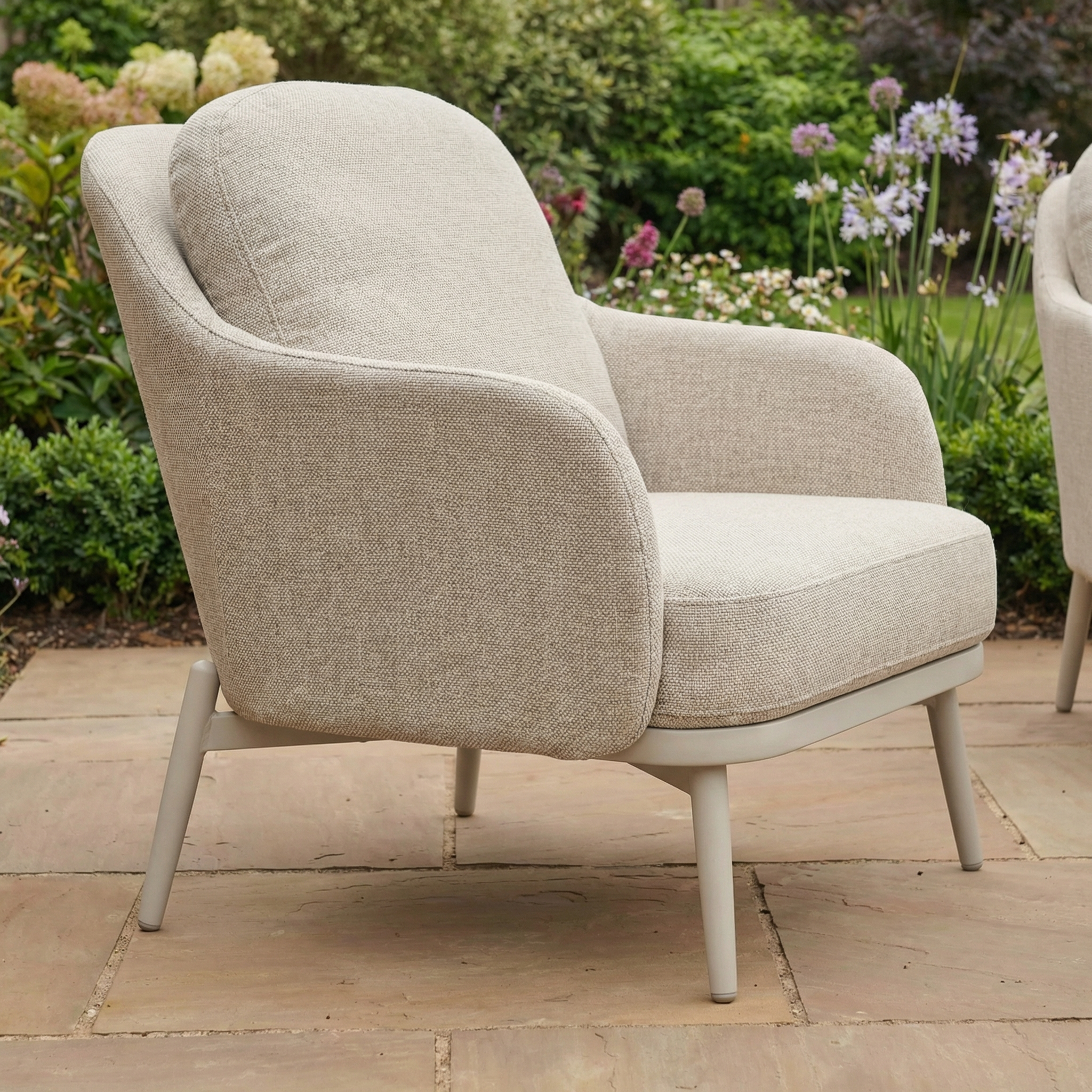 A beige cushioned armchair with wooden legs on a stone patio complements outdoor pieces like the Alva 3 Seat Sofa Dining Set with Rising Table in Fawn, creating a stylish and relaxing garden retreat.
