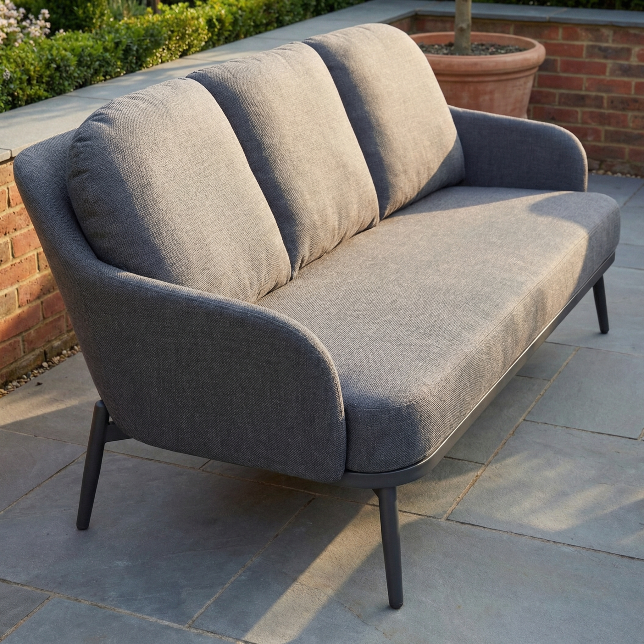 The Alva 3 Seat Sofa Dining Set with Rising Table in Grey features stylish cushioned seating—perfect garden furniture for relaxing outdoors, shown here on a stone patio by a brick wall and potted plant.