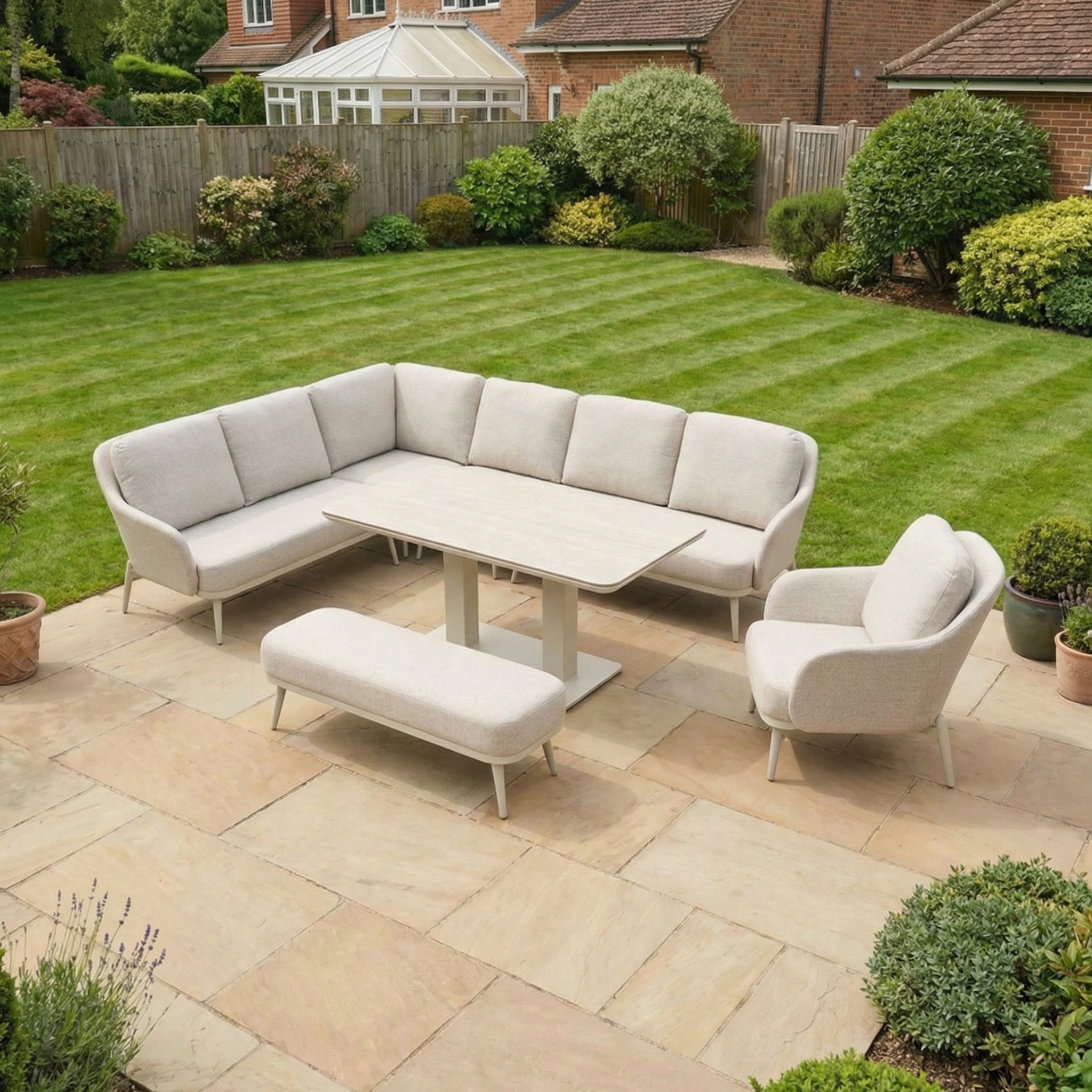 The Alva Corner Dining Set with Rising Table and Armchair in Fawn offers elegant garden furniture, featuring cream cushions on a stone terrace overlooking a green lawn and garden for style and convenience.
