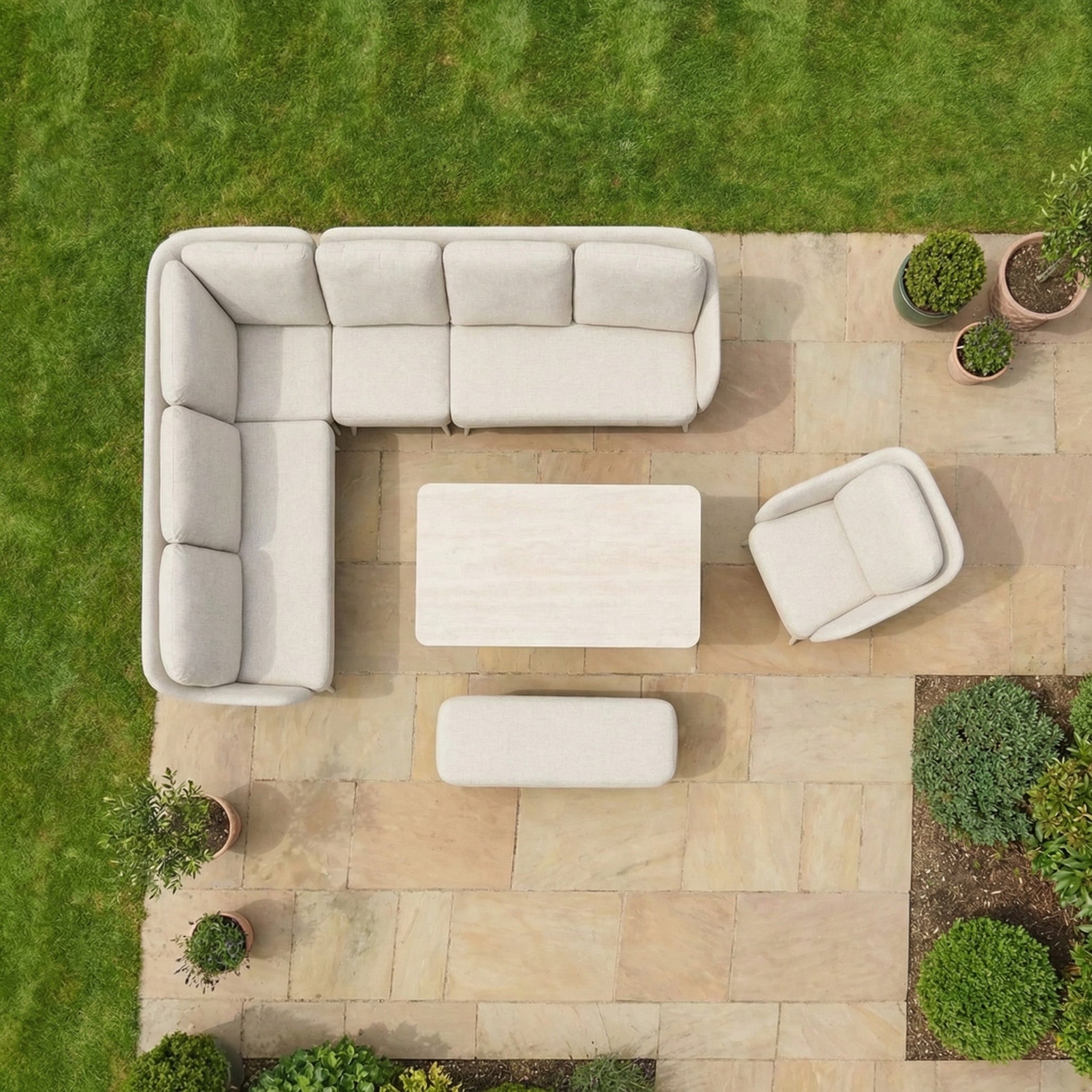 The Alva Corner Dining Set with Rising Table and Armchair in Fawn, placed on a stone patio amid green grass and potted plants, creates a perfect spot for stylish garden furniture.