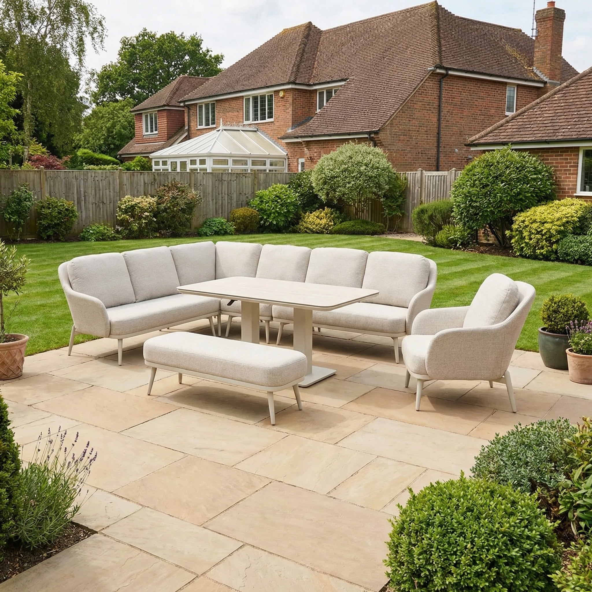 Outdoor patio featuring the Alva Corner Dining Set with Rising Table and Armchair in Fawn, plus bench seating, overlooking a garden and large brick house.