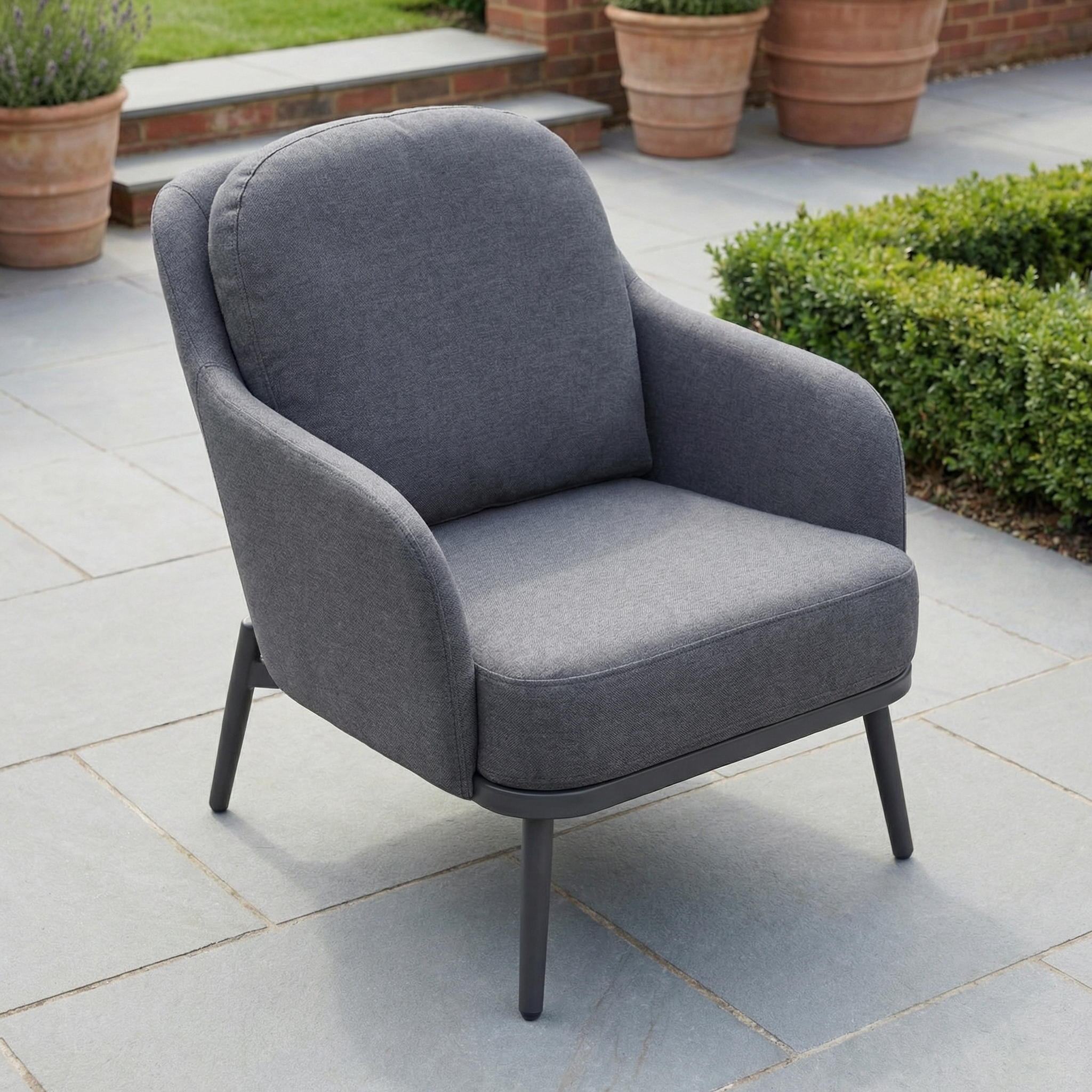 The Alva Corner Dining Set with Rising Table and Armchair in Grey (Right Hand) is arranged on a stone patio, surrounded by potted plants, green hedges, and stylish garden furniture.