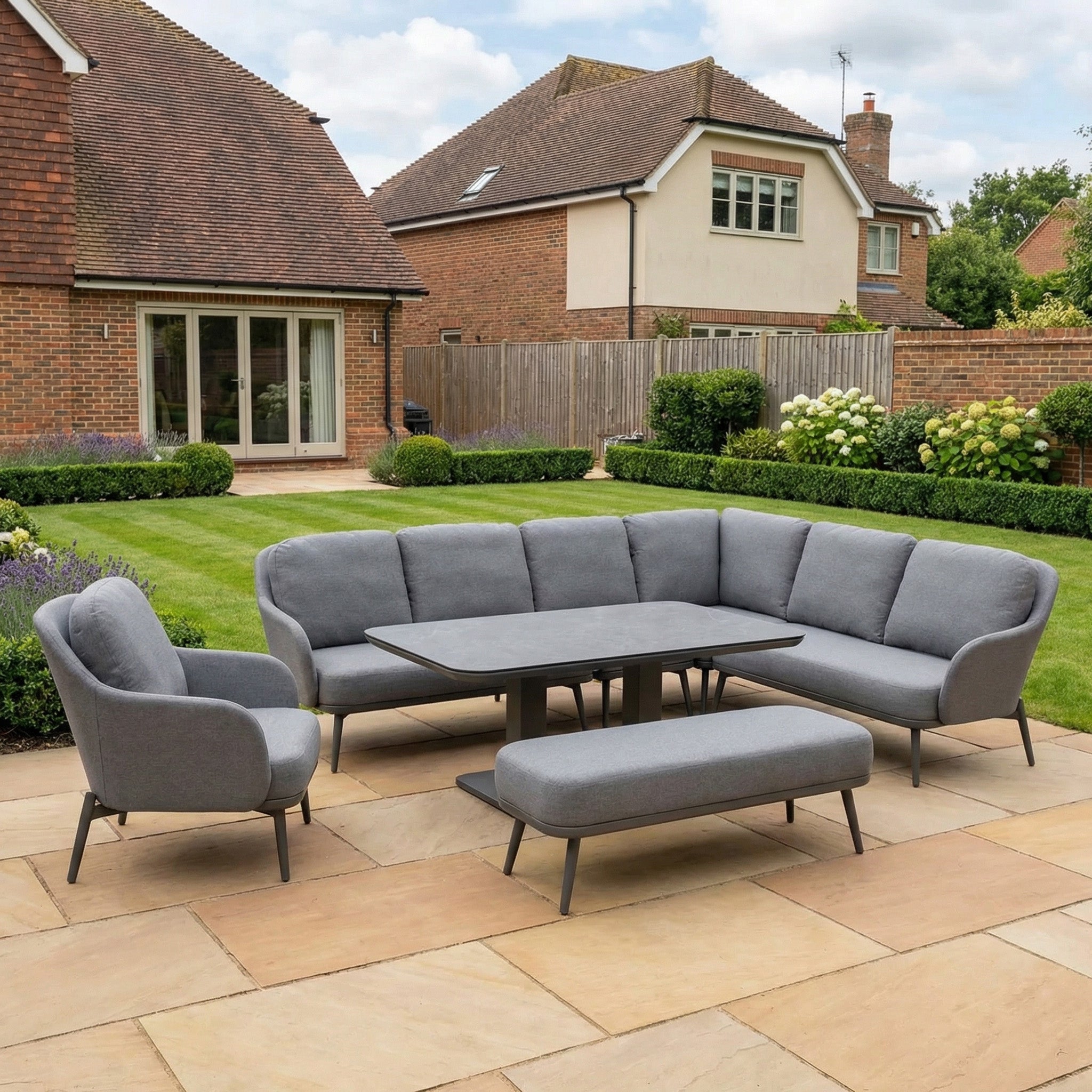 The Alva Corner Dining Set with Rising Table and Armchair in Grey (Right Hand) sits on a stone patio between two brick houses, offering stylish and versatile outdoor entertaining in your landscaped backyard.
