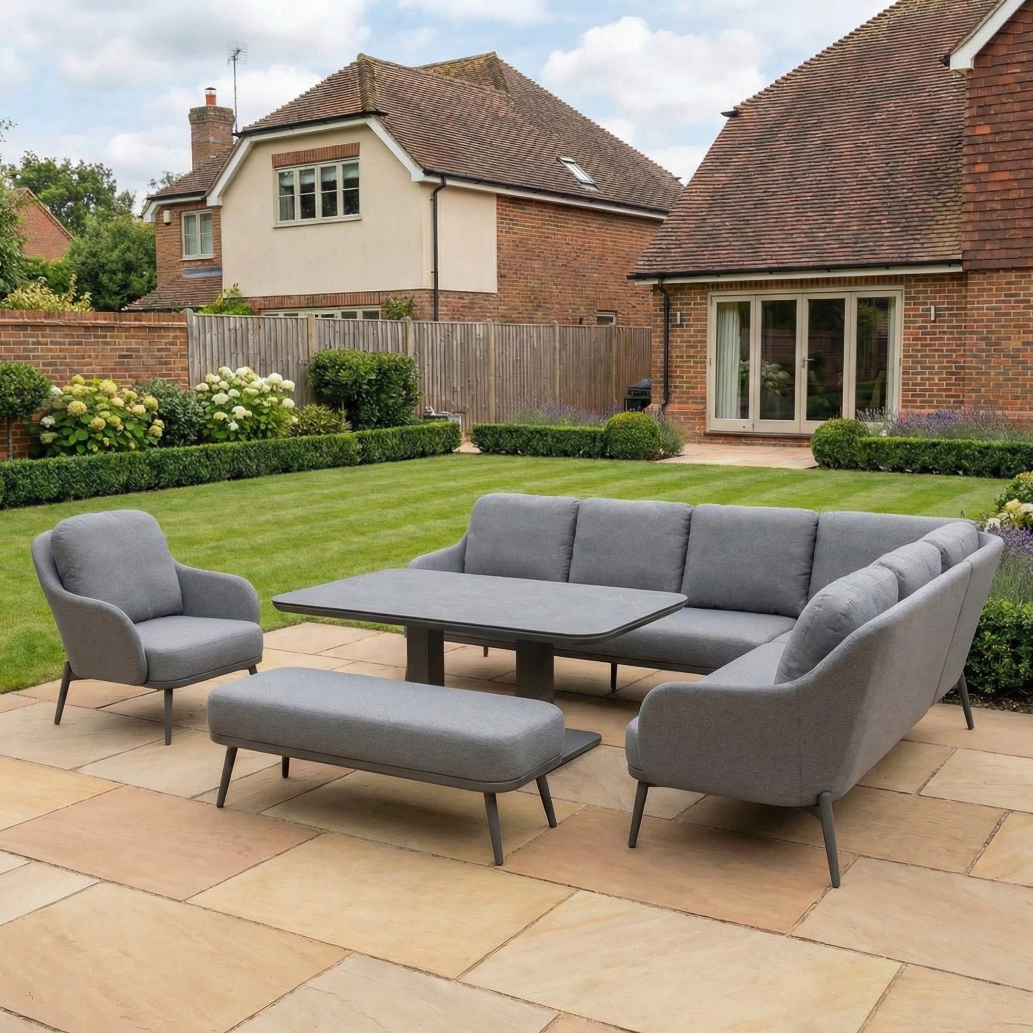 The Alva Corner Dining Set with Rising Table and Armchair in Grey (Right Hand) is shown on a tiled patio in a backyard with grass, shrubs, and brick houses in the background.
