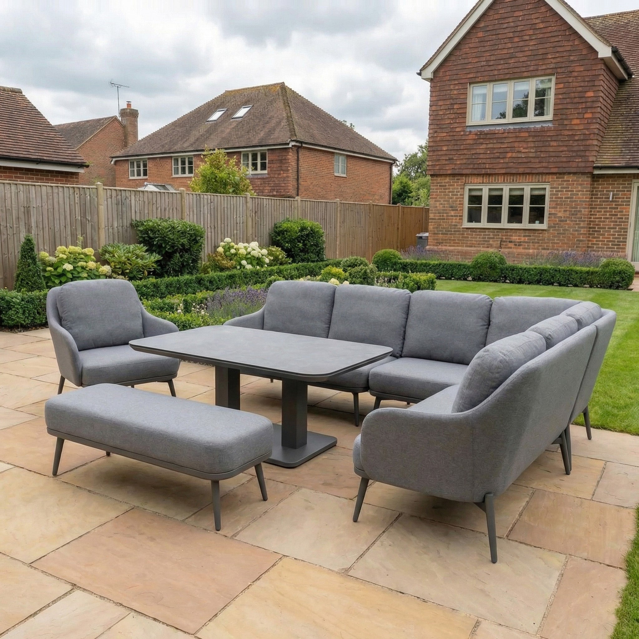 The Alva Corner Dining Set with Rising Table and Armchair in Grey (Right Hand) sits on a stone terrace, offering modern style and flexibility, with houses and a lush green garden as the scenic backdrop.