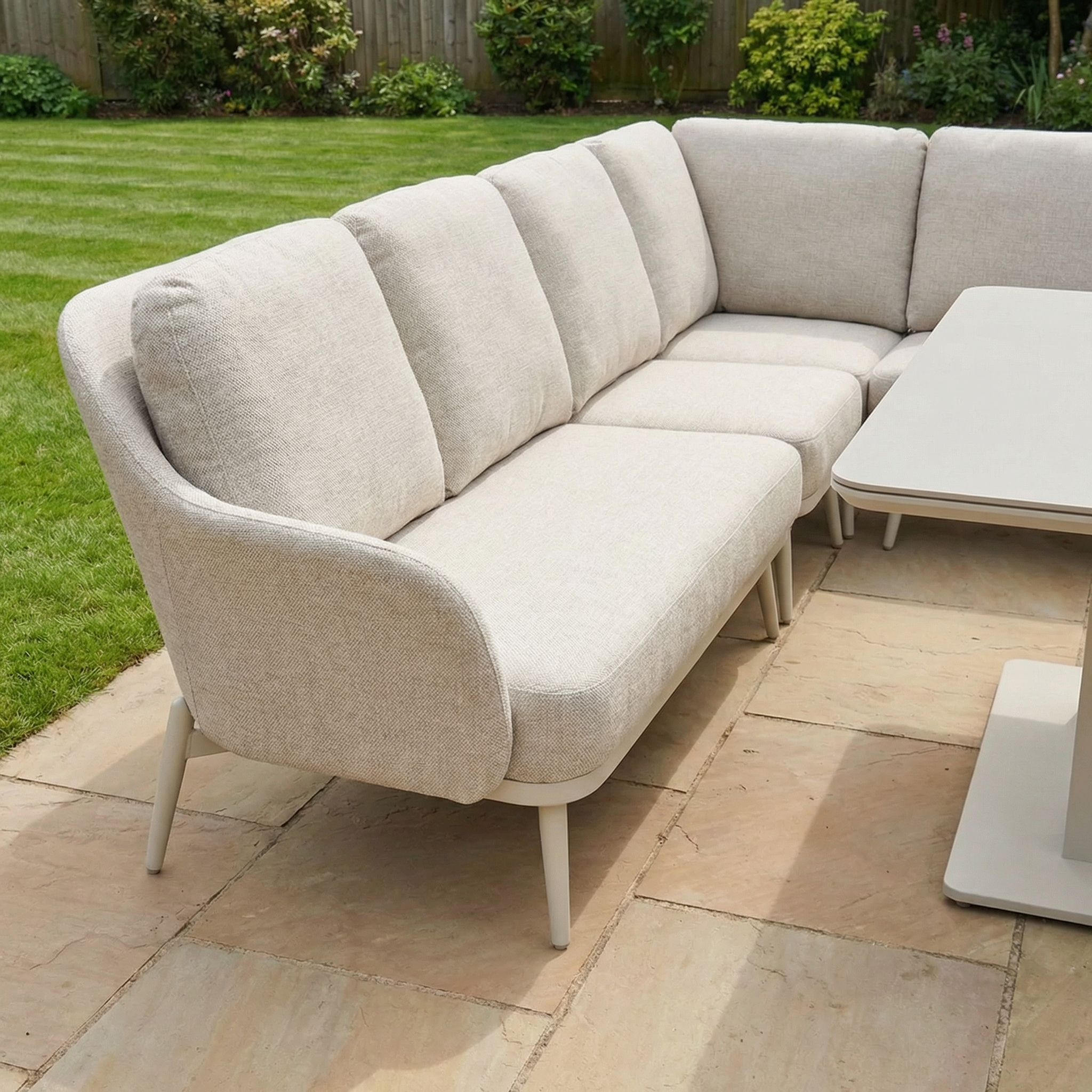 The Alva Corner Dining Set with Rising Table in Fawn (Right Hand) sits on a stone patio by a lawn and shrubs, offering garden comfort and a versatile rising table for convenient outdoor dining.