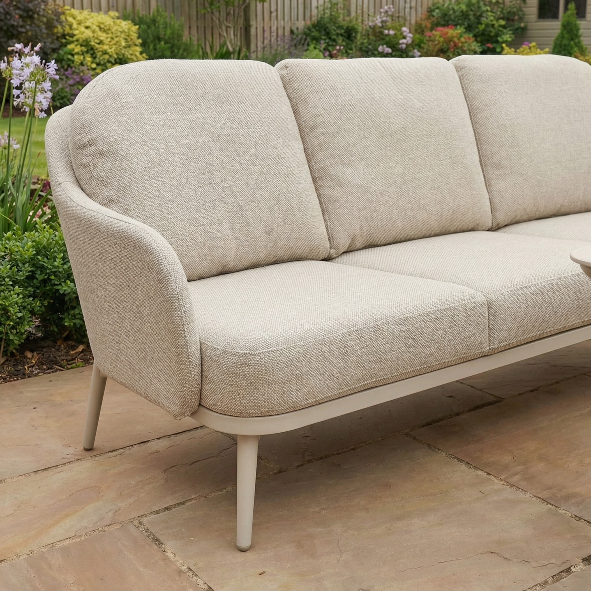 Relax or entertain outdoors with the Alva Corner Dining Set with Rising Table in Fawn (Right Hand)—a beige outdoor sofa featuring soft cushions, set on a stone patio and surrounded by a lush green garden.