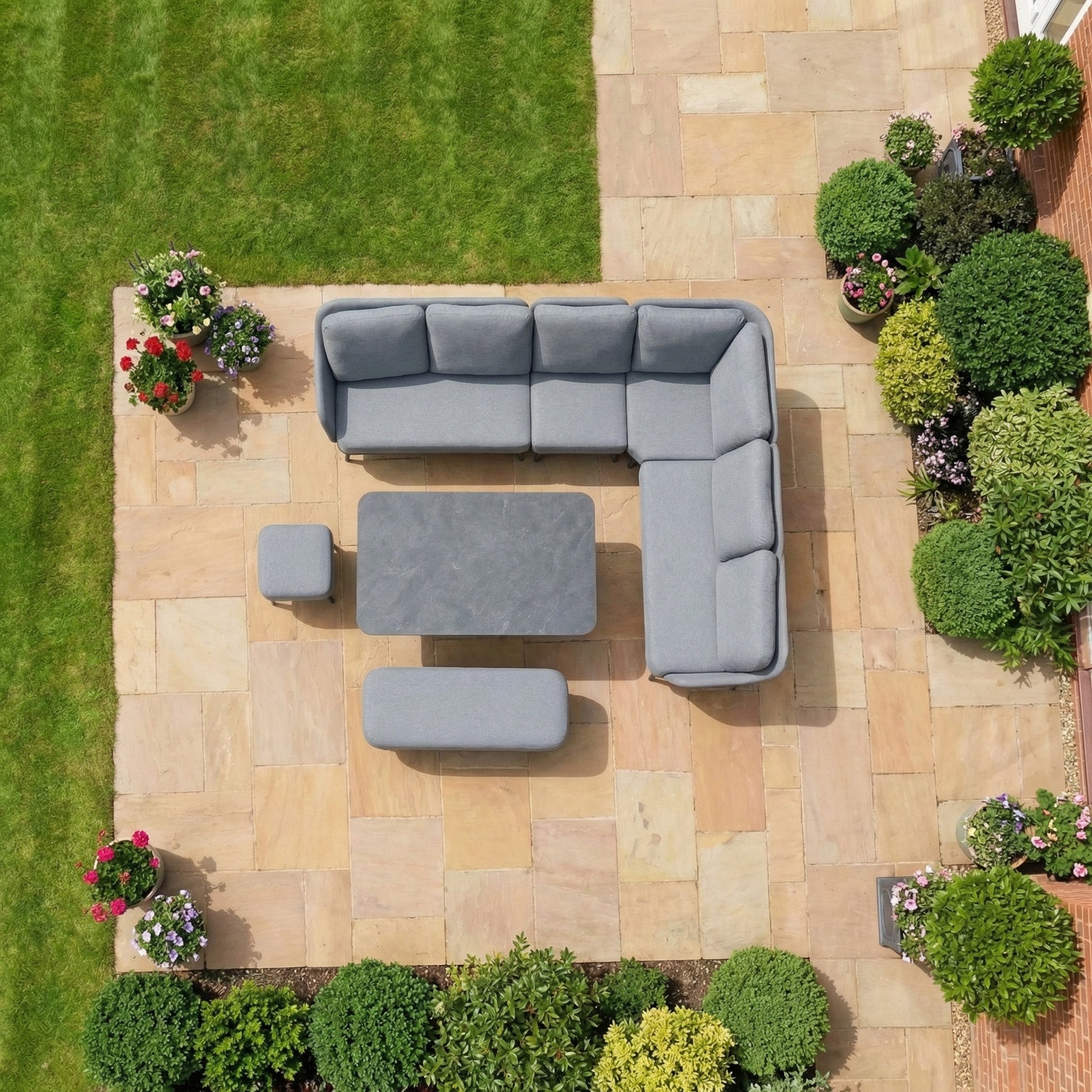The Alva Corner Dining Set with Rising Table in Grey includes a gray outdoor sectional sofa, ottomans, and a versatile rising table, perfect for stone patios surrounded by green lawns and garden furniture.