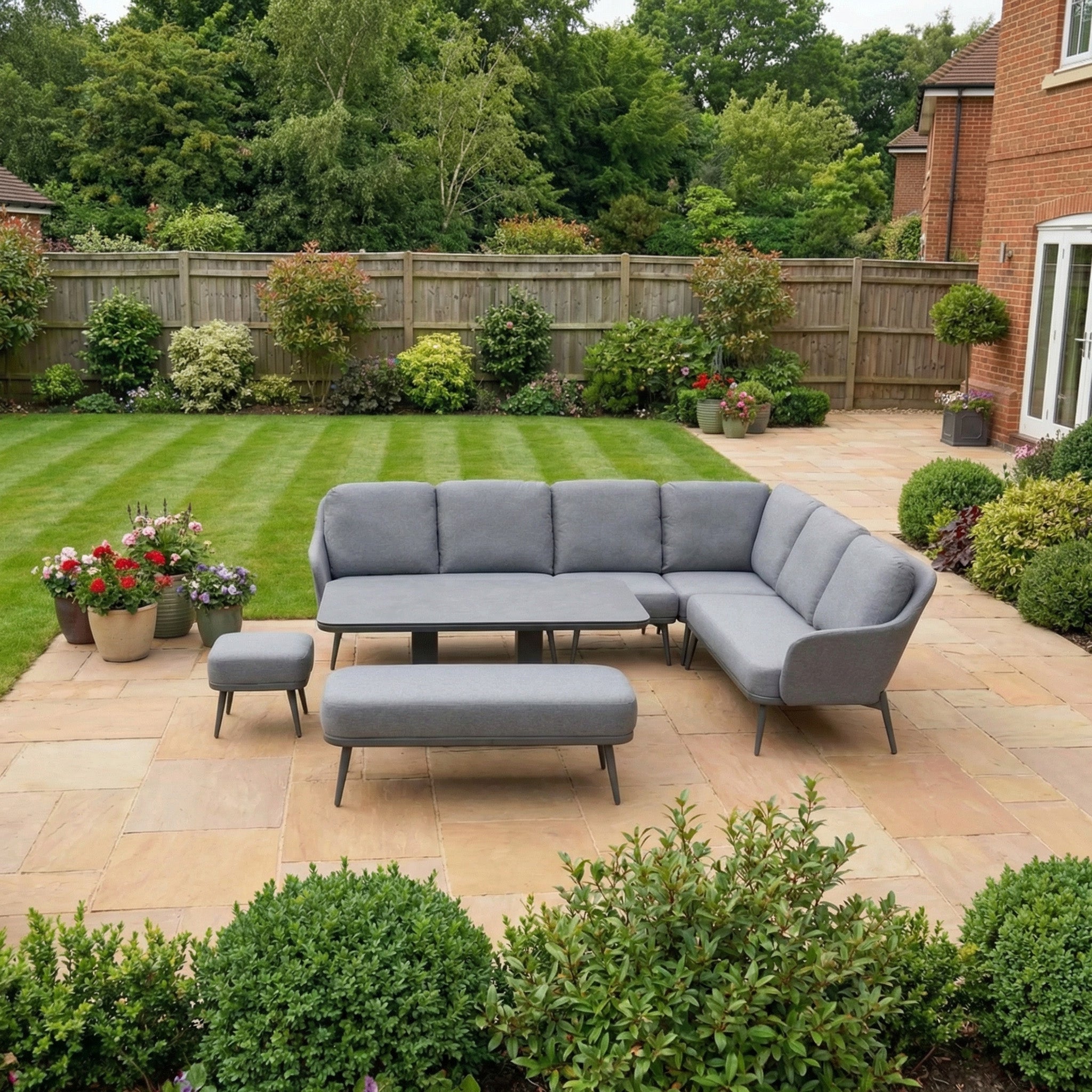 The Alva Corner Dining Set with Rising Table in Grey, featuring benches, is arranged on a stone patio bordered by lawn, plants, and a wooden fence.