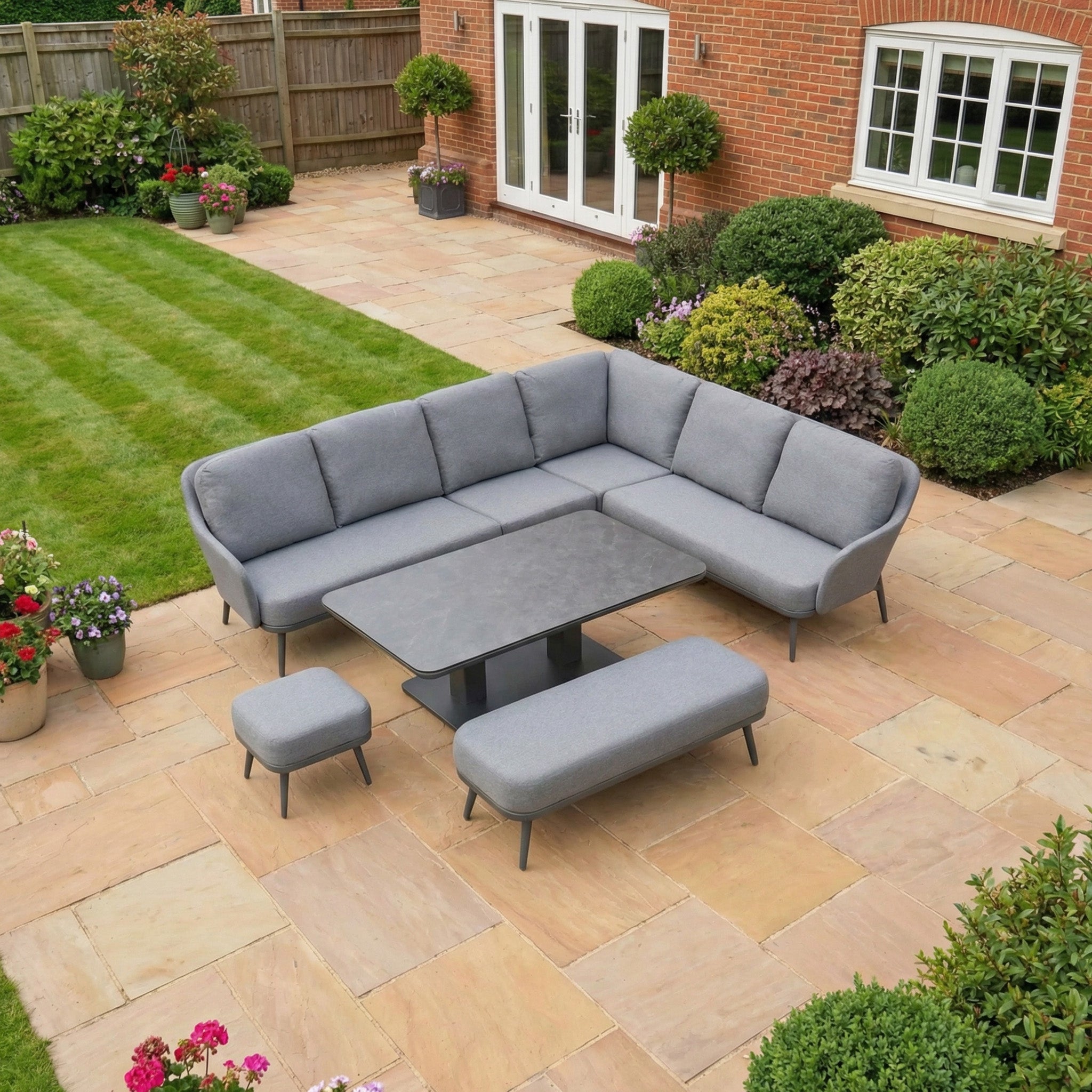 The Alva Corner Dining Set with Rising Table in Grey includes a sectional sofa, bench, stool, and multi-use table—ideal for outdoor spaces with a stone patio, greenery, and lawn.