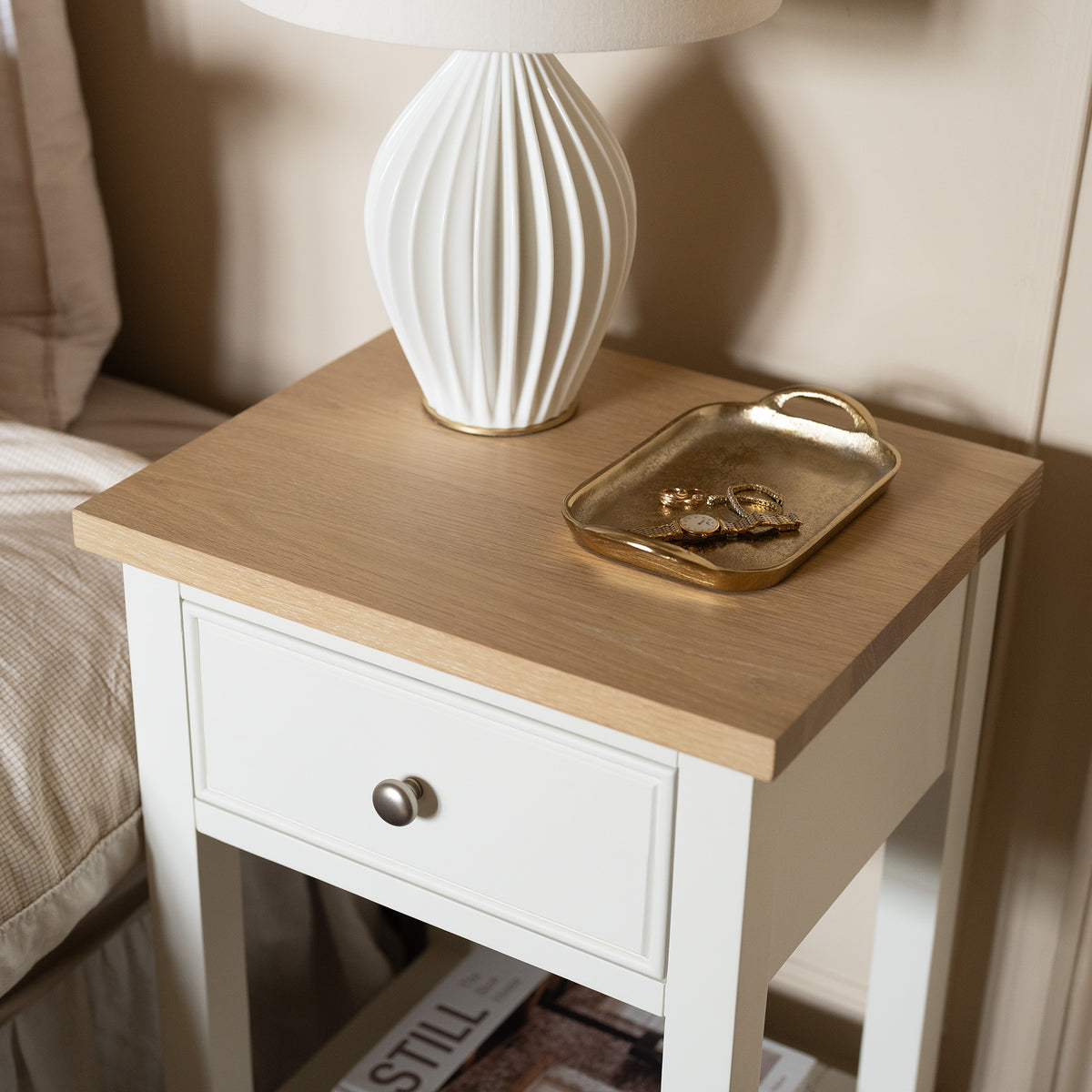 Amberley Bedside Table in Warm White, styled with a lamp and a gold tray for keys, placed beside a bed with a magazine on the shelf below, adds modern country furniture charm to your space.