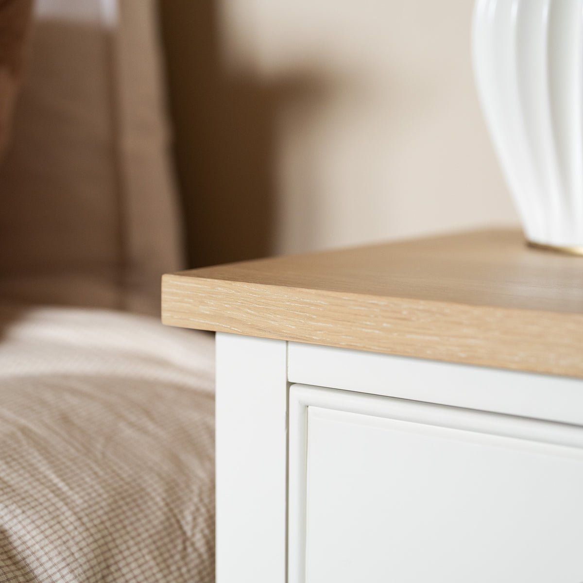 Close-up of the Amberley Bedside Table in Warm White, featuring a light wood top and styled in modern country fashion, placed beside a bed and a white lamp base. This bedside table beautifully blends functionality with timeless charm, making it an ideal addition to any bedroom setting.