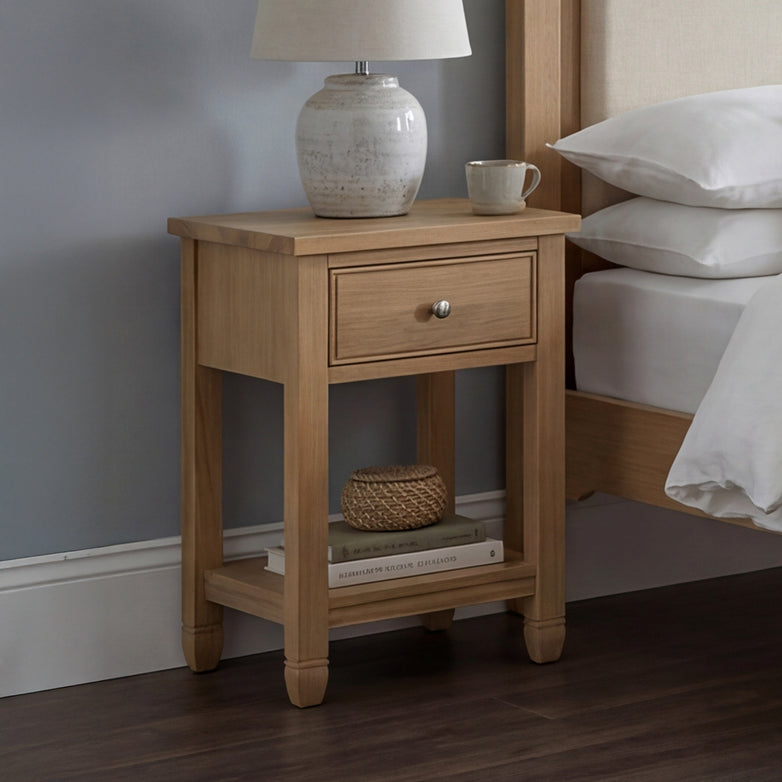 The Amberley Bedside Table in Light Oak sits by the bed, holding a lamp, mug, basket, and books against a blue wall.