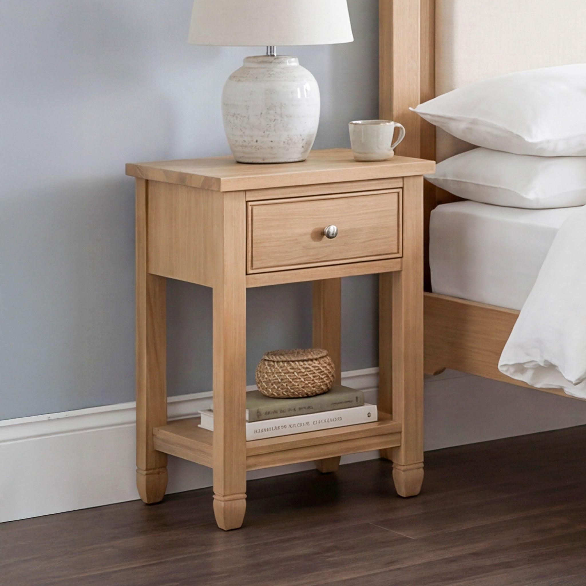 The Amberley Bedside Table in Light Oak, topped with a lamp, mug, small basket, and books, stands beside a bed on a dark wood floor.