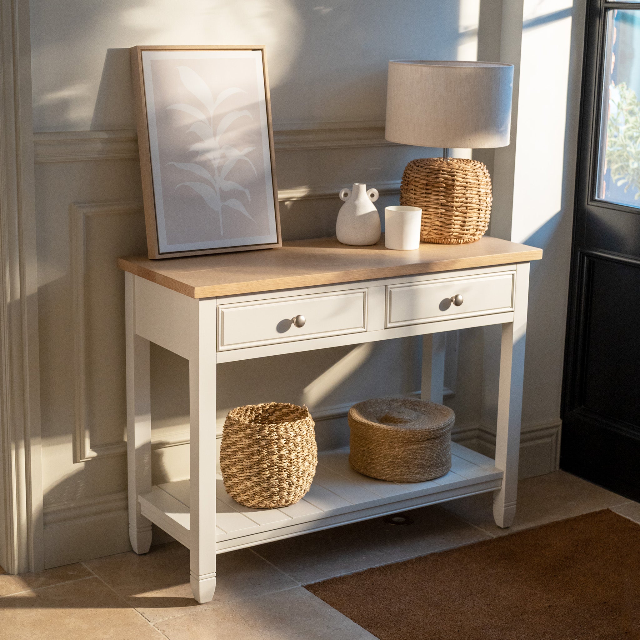 The Amberley 2 Drawers Console Table in Warm White, featuring an oak top, is styled with a lamp, framed art, and woven baskets in a sunlit entryway.