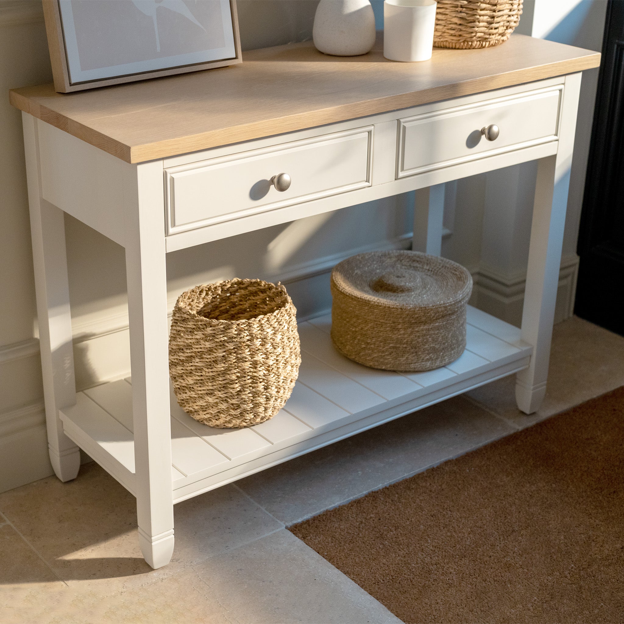 The Amberley 2 Drawers Console Table in Warm White features an oak top and woven baskets on the lower shelf, adding farmhouse style to any sunlit room.