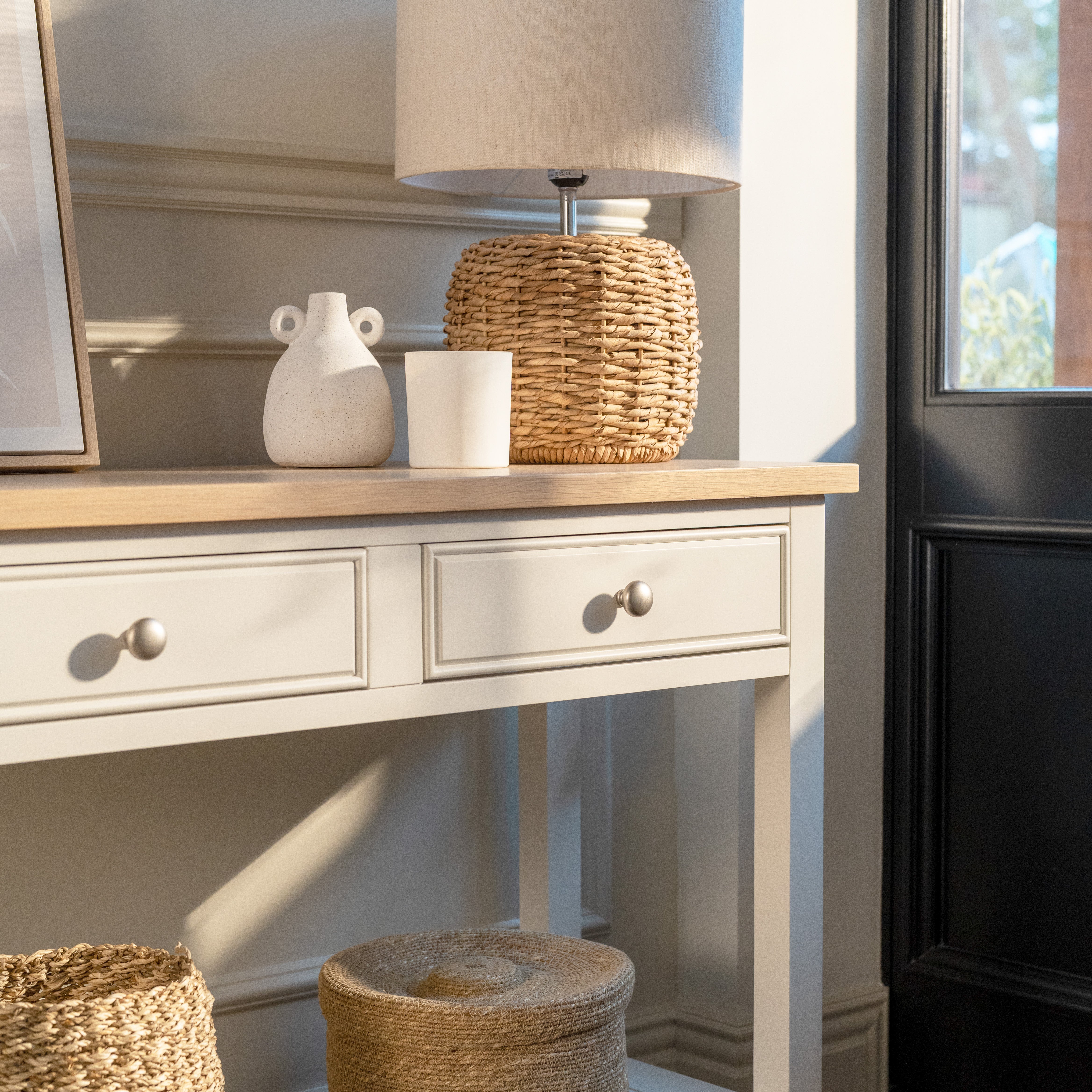 The Amberley 2 Drawers Console Table in Warm White, topped with a lamp, vases, and basket by the door in a sunlit room, brings subtle farmhouse charm.