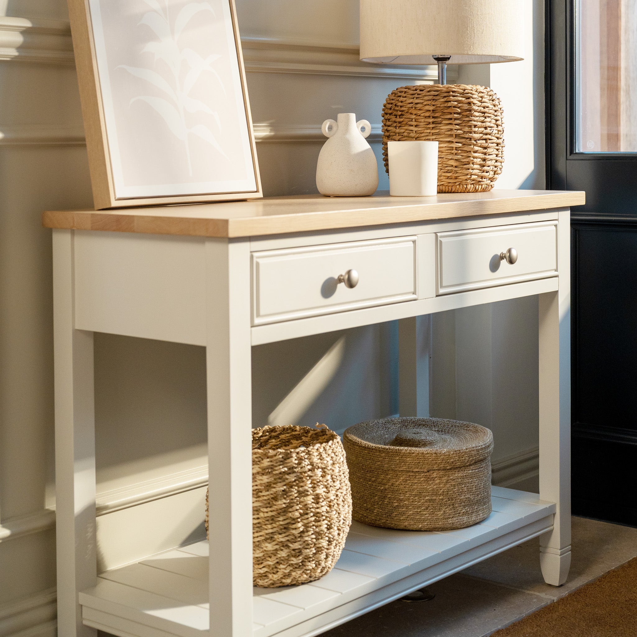 The Amberley 2 Drawers Console Table in Warm White, shown with wicker baskets, a lamp, and decor near a door, features an oak top that adds warmth and charm to farmhouse-style spaces filled with bright natural light.