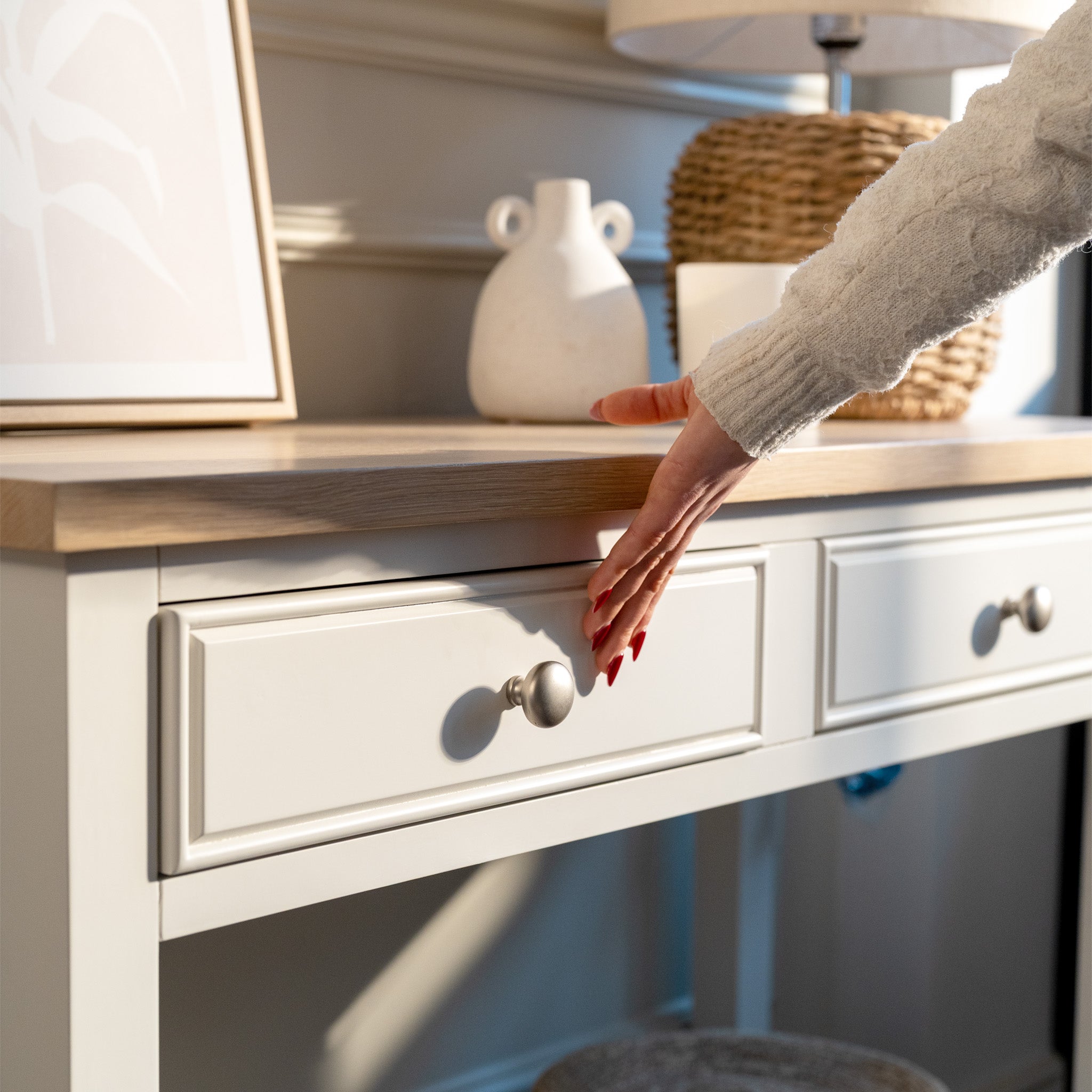 A person’s hand reaches to open a drawer on the Amberley 2 Drawers Console Table in Warm White, featuring an oak top and farmhouse style, in a sunlit room.