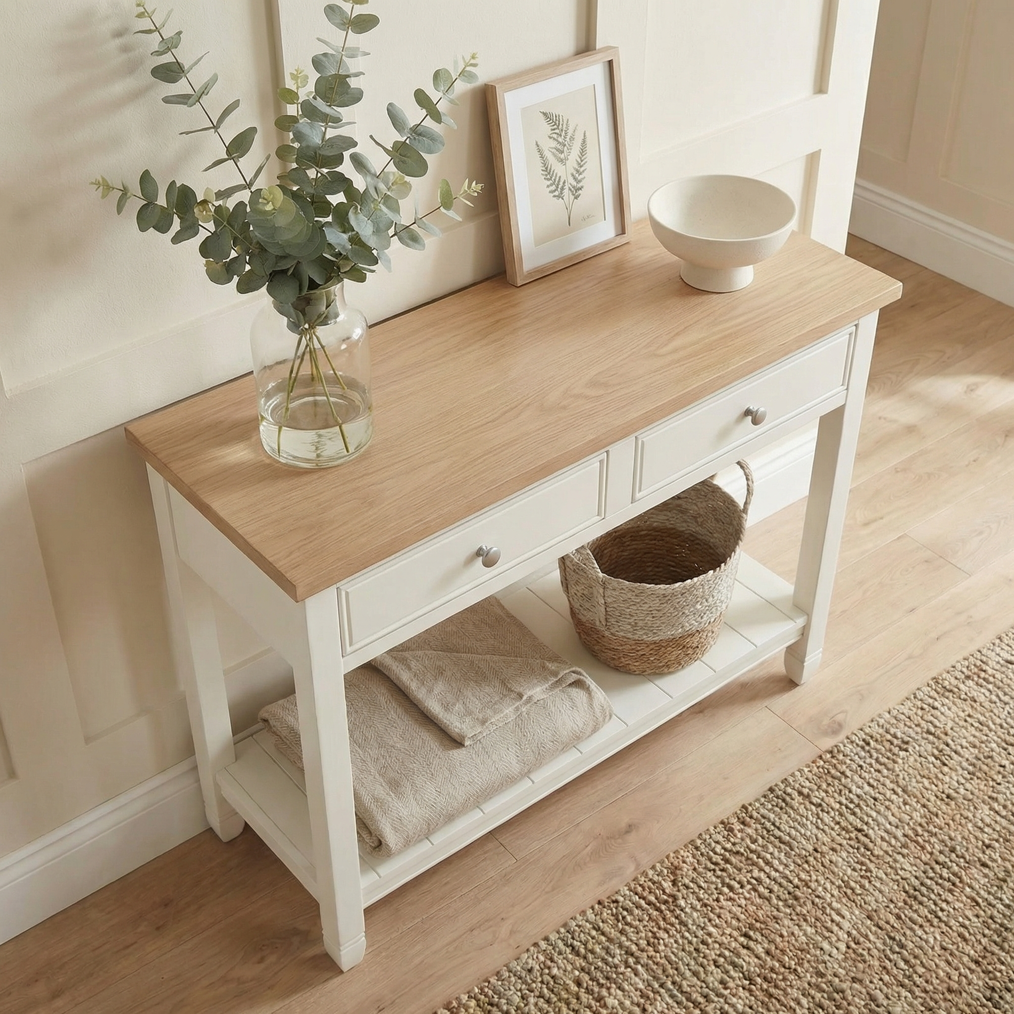The Amberley 2 Drawers Console Table in Warm White features an oak top and farmhouse style, with drawers, plus space for decor like a vase, framed art, bowl, basket, and folded towels on the shelf.