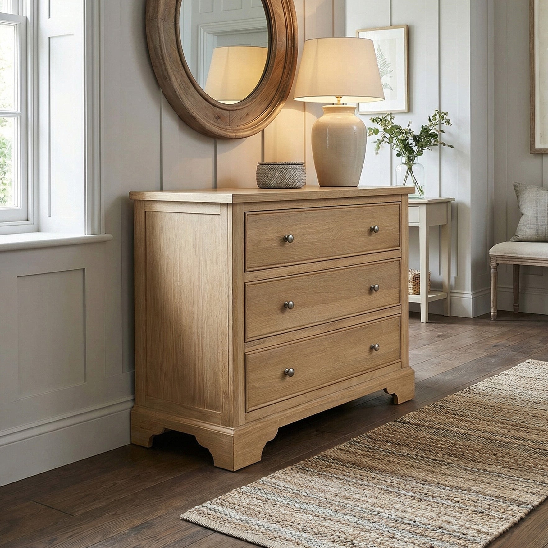 The Amberley 3 Chest of Drawers in Light Oak, topped with a lamp and decor, stands in a bright, cozy room beneath a round mirror.