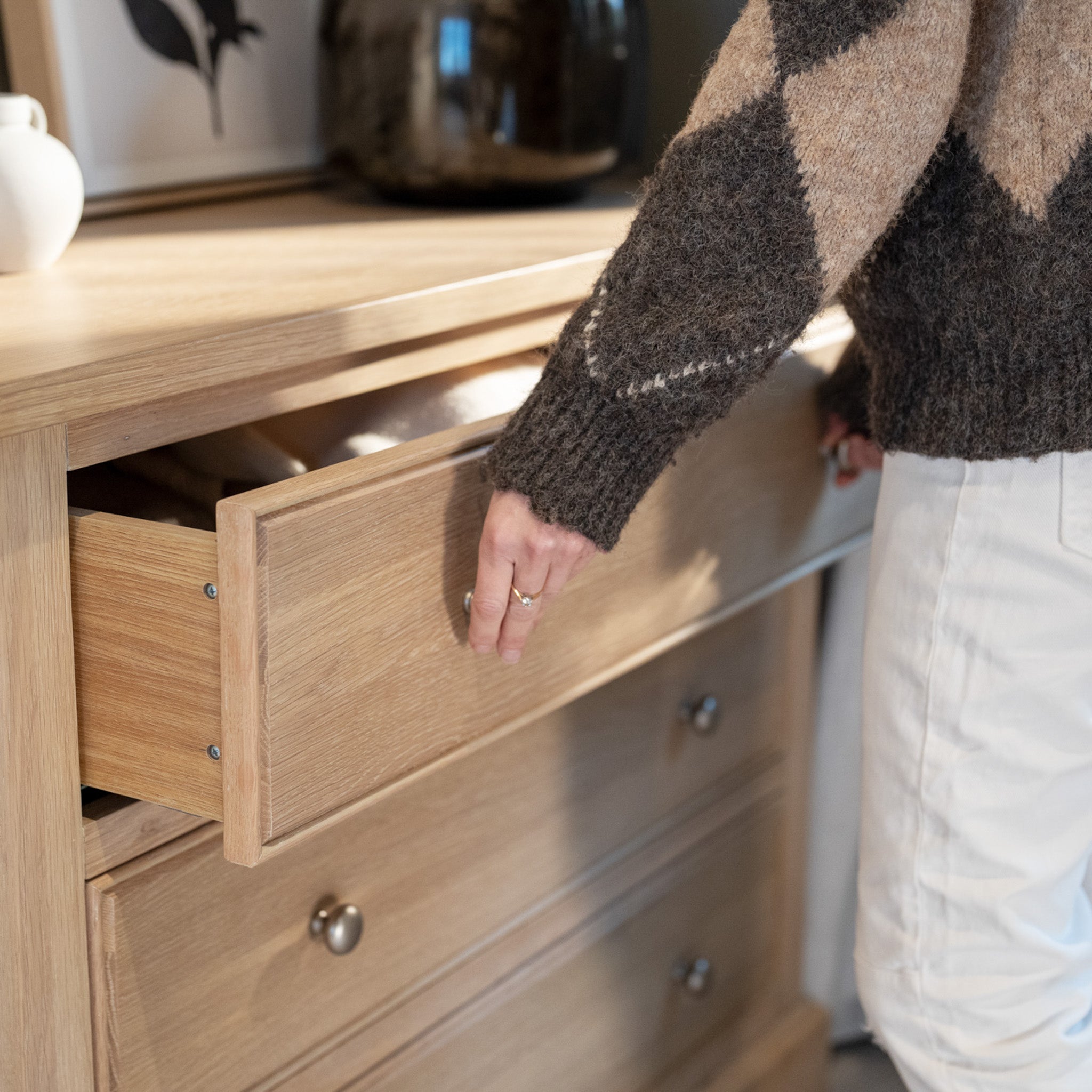 Wearing a patterned sweater and light-colored pants, a person opens the Amberley 3 Chest of Drawers in Light Oak.