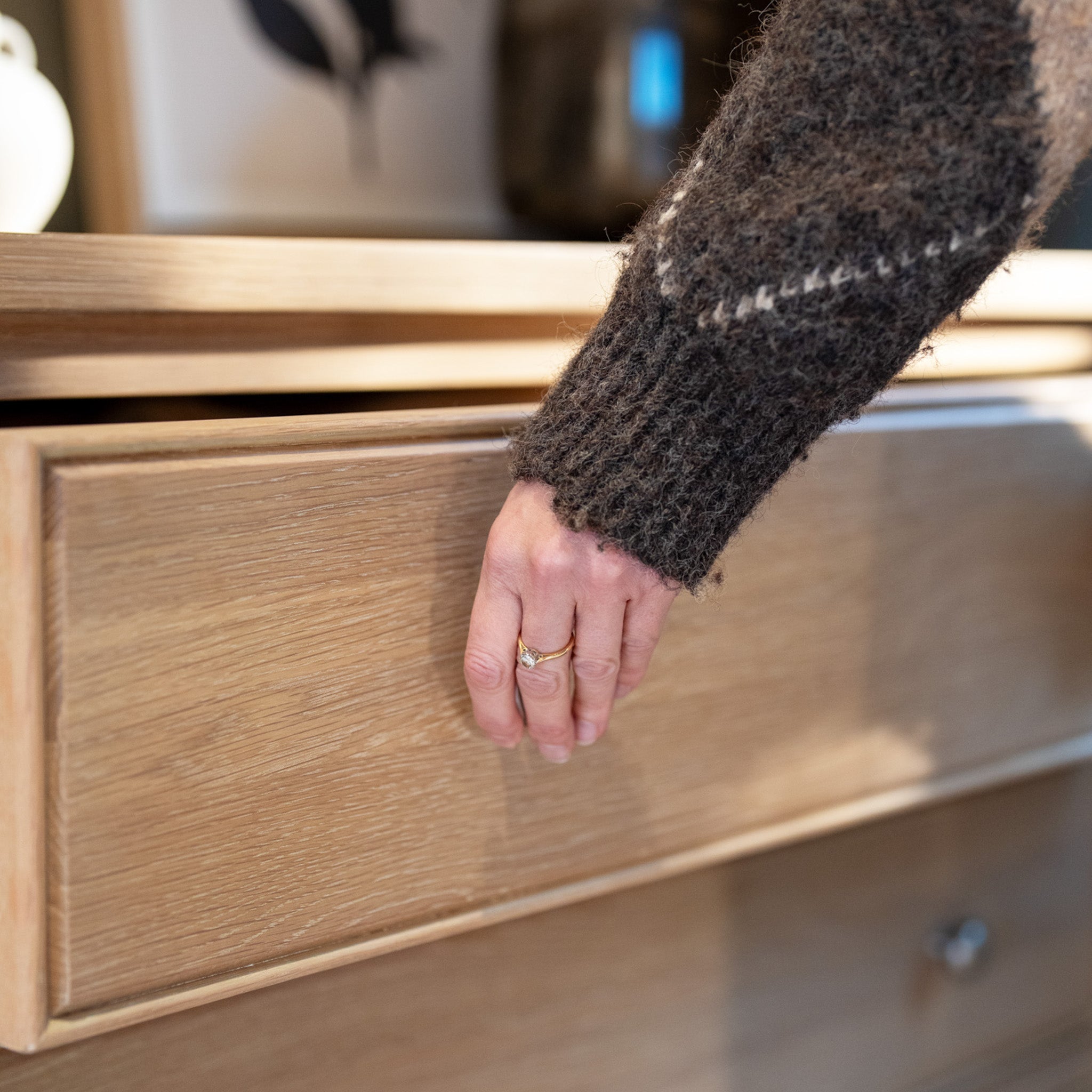 A person wearing a wool sweater opens the wooden drawer of the Amberley 3 Chest of Drawers in Light Oak with their left hand.