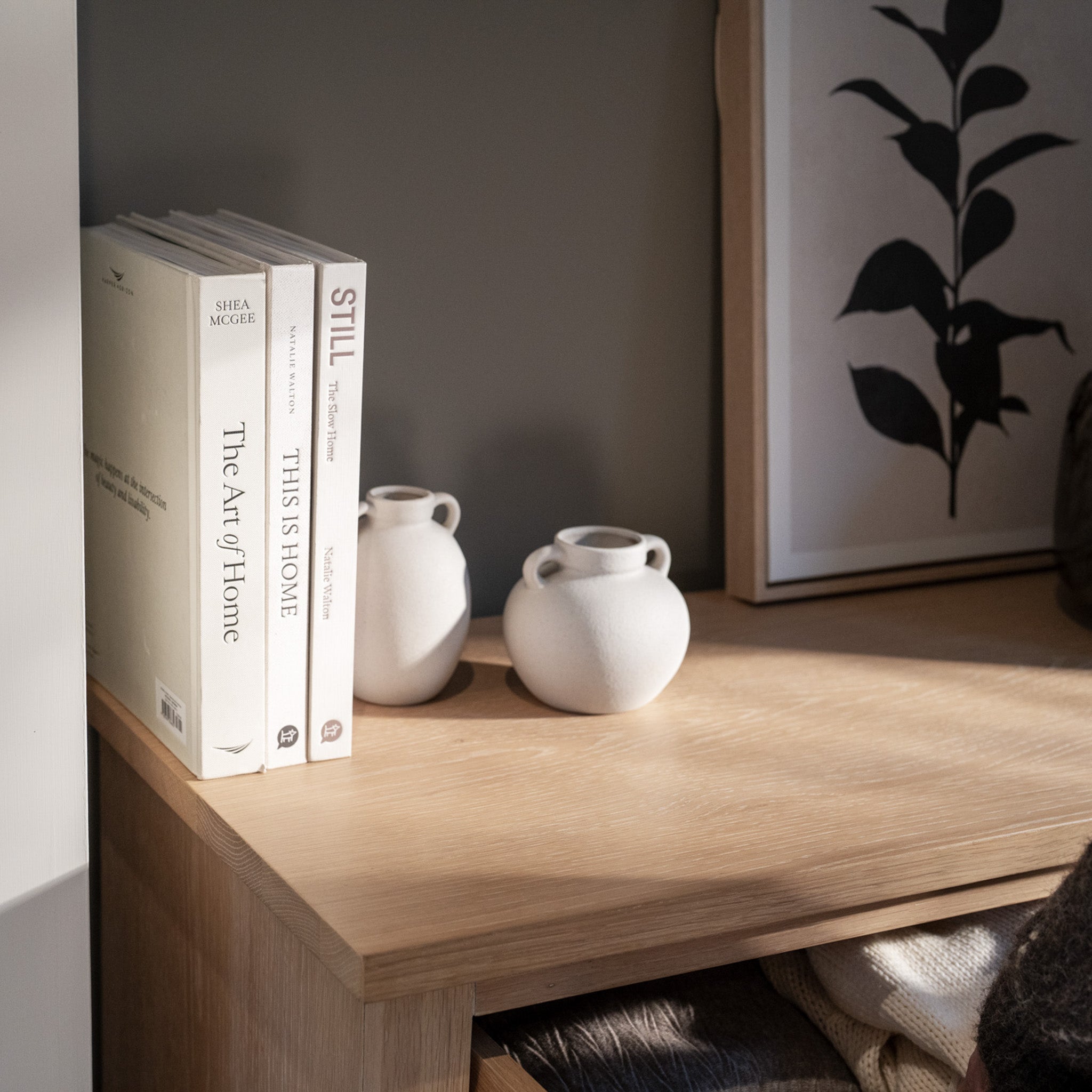 Books and small white vases sit on a wooden shelf before a botanical art print, beautifully complementing the nearby Amberley 3 Chest of Drawers in Light Oak.