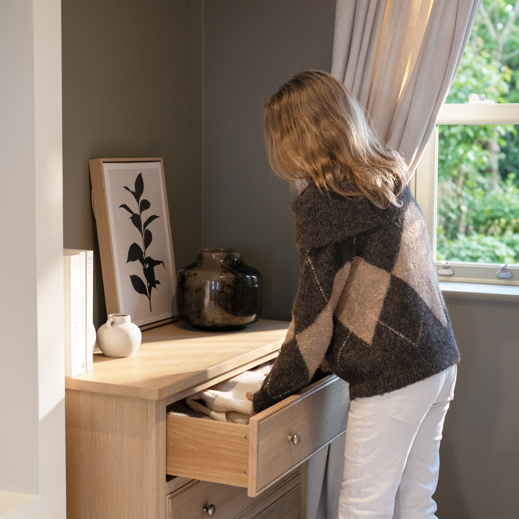 A woman in a sweater opens a drawer of the Amberley 3 Chest of Drawers in Light Oak, placed in a cozy, sunlit room decorated with items on top.