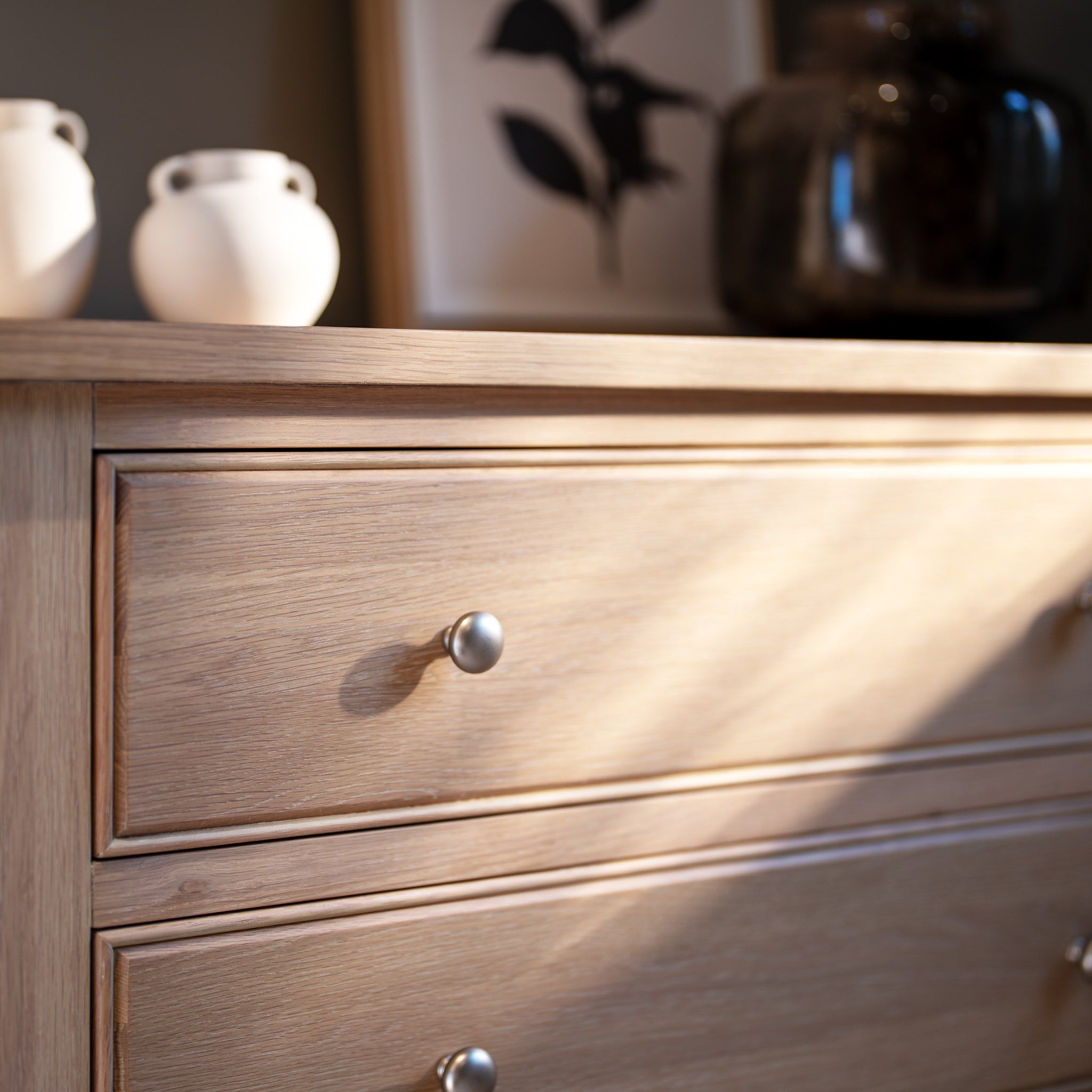 Amberley 3 Chest of Drawers in Light Oak with silver knobs, styled with vases and framed art in the background.